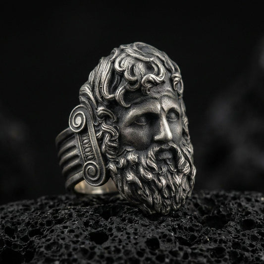 Detailed Zeus ring crafted in oxidized sterling silver, classical Greek god face with flowing beard and ornate curls, macro product shot on textured volcanic rock background