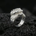 Load image into Gallery viewer, Polished silver Foo Dog ring featuring sculpted guardian lion face and flowing scrollwork details, centered close-up on dark porous volcanic stone
