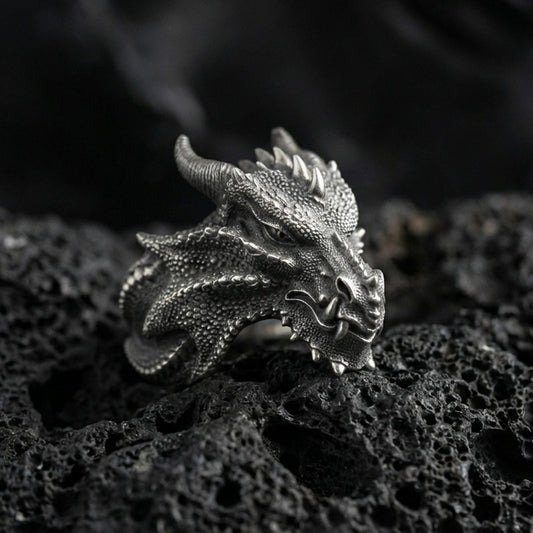 Detailed dragon wyrm ring crafted in oxidized sterling silver, textured scale pattern with horned dragon head, macro product shot on textured volcanic rock background