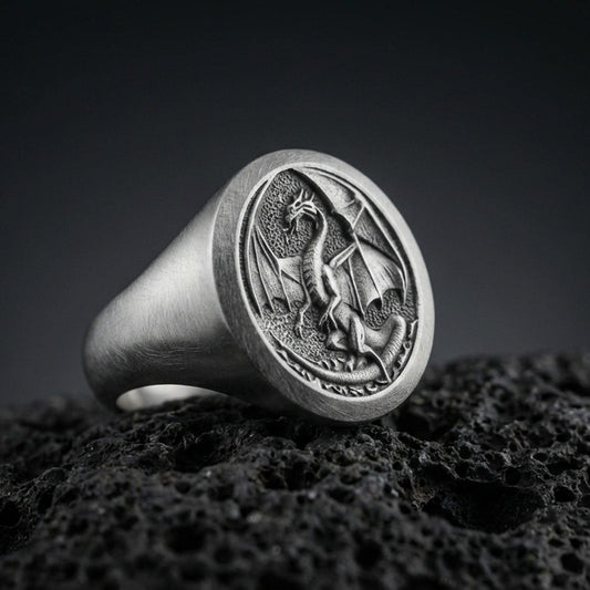 Detailed Chinese dragon signet ring crafted in oxidized sterling silver, coiled dragon with spread wings relief, macro product shot on textured volcanic rock background