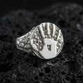 Load image into Gallery viewer, Polished silver handprint seal ring featuring raised palm emblem and micro skull side details, macro shot on porous volcanic stone
