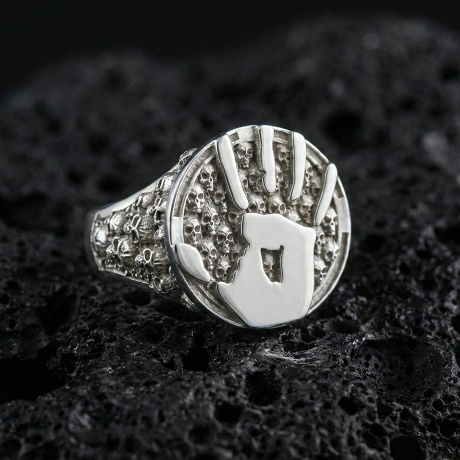 Polished silver handprint seal ring featuring raised palm emblem and micro skull side details, macro shot on porous volcanic stone