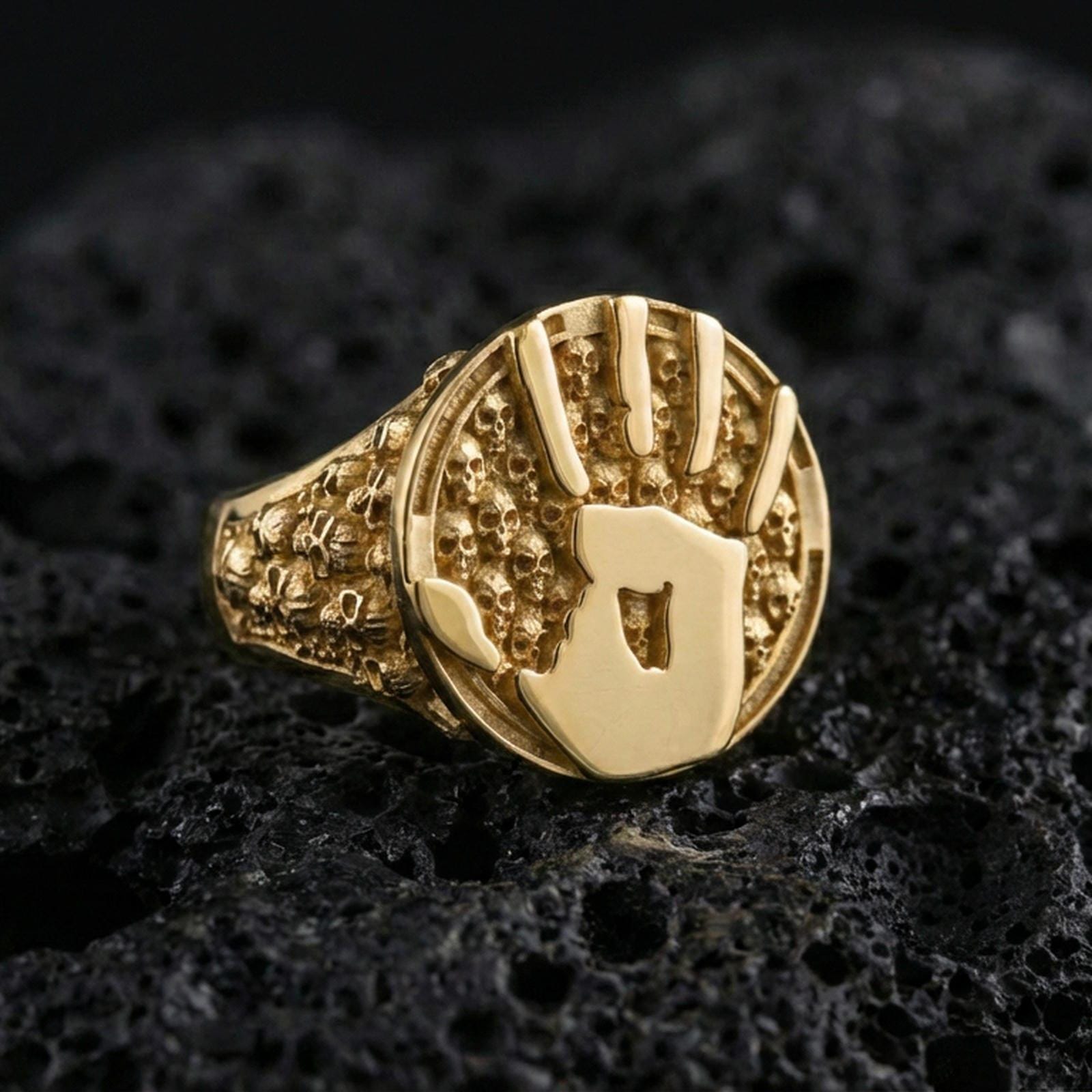 Gold plated handprint signet ring with gothic skull band and round textured face, premium studio close-up on volcanic rock background