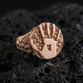 Load image into Gallery viewer, Rose gold plated assassin emblem ring with handprint motif and skull-pattern band, luxury macro product photo on volcanic rock surface

