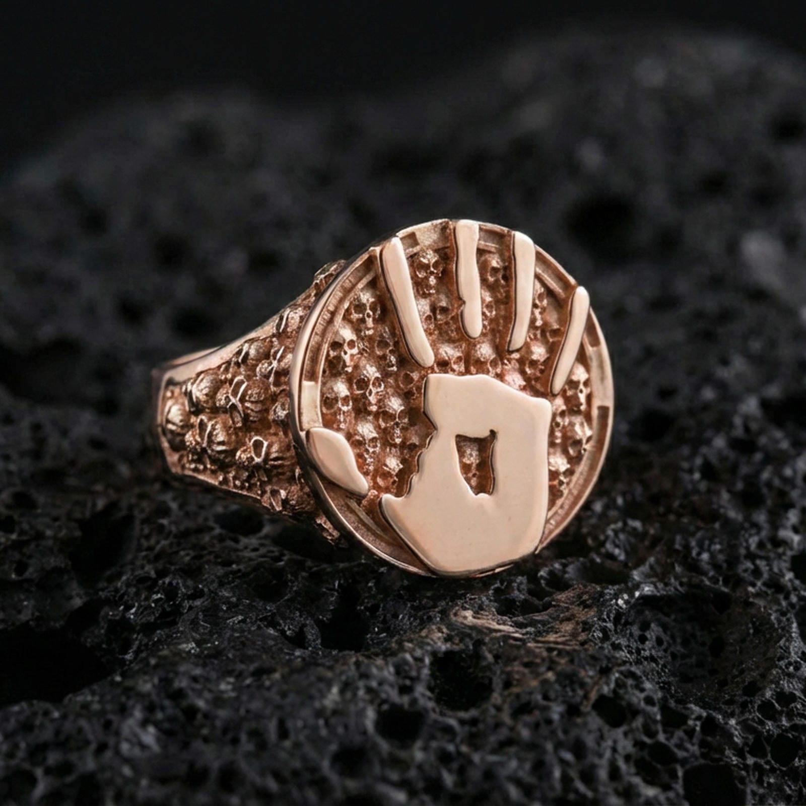 Rose gold plated assassin emblem ring with handprint motif and skull-pattern band, luxury macro product photo on volcanic rock surface