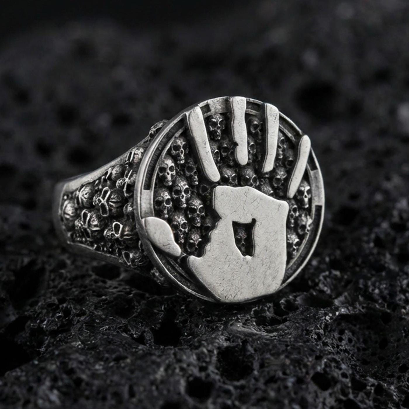 Oxidized sterling silver assassin handprint signet ring with skull band texture, dark fantasy statement jewelry on black volcanic rock
