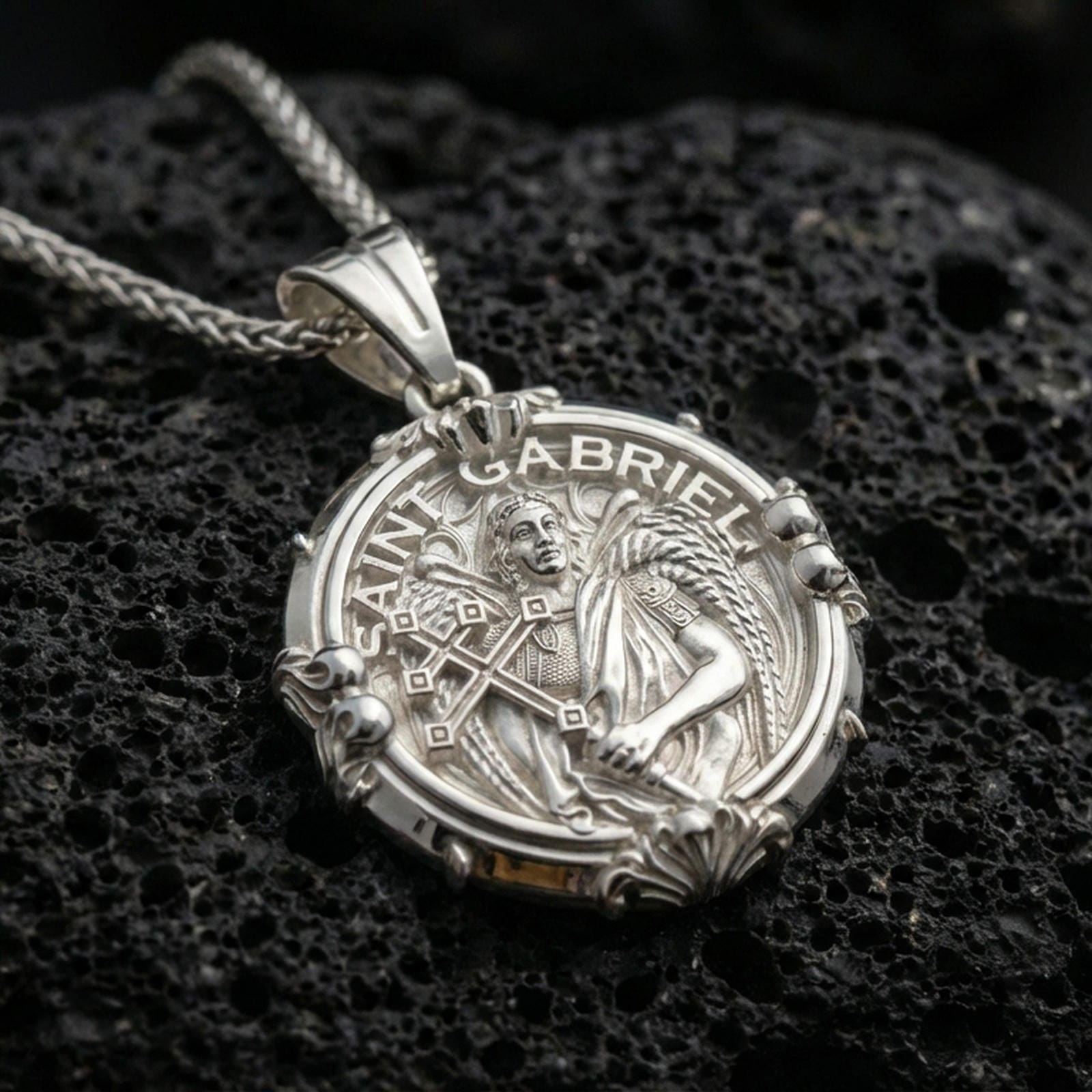 Polished silver Saint Gabriel medallion necklace on dark volcanic stone, crisp angel figure detail and ornamental border, close-up shot