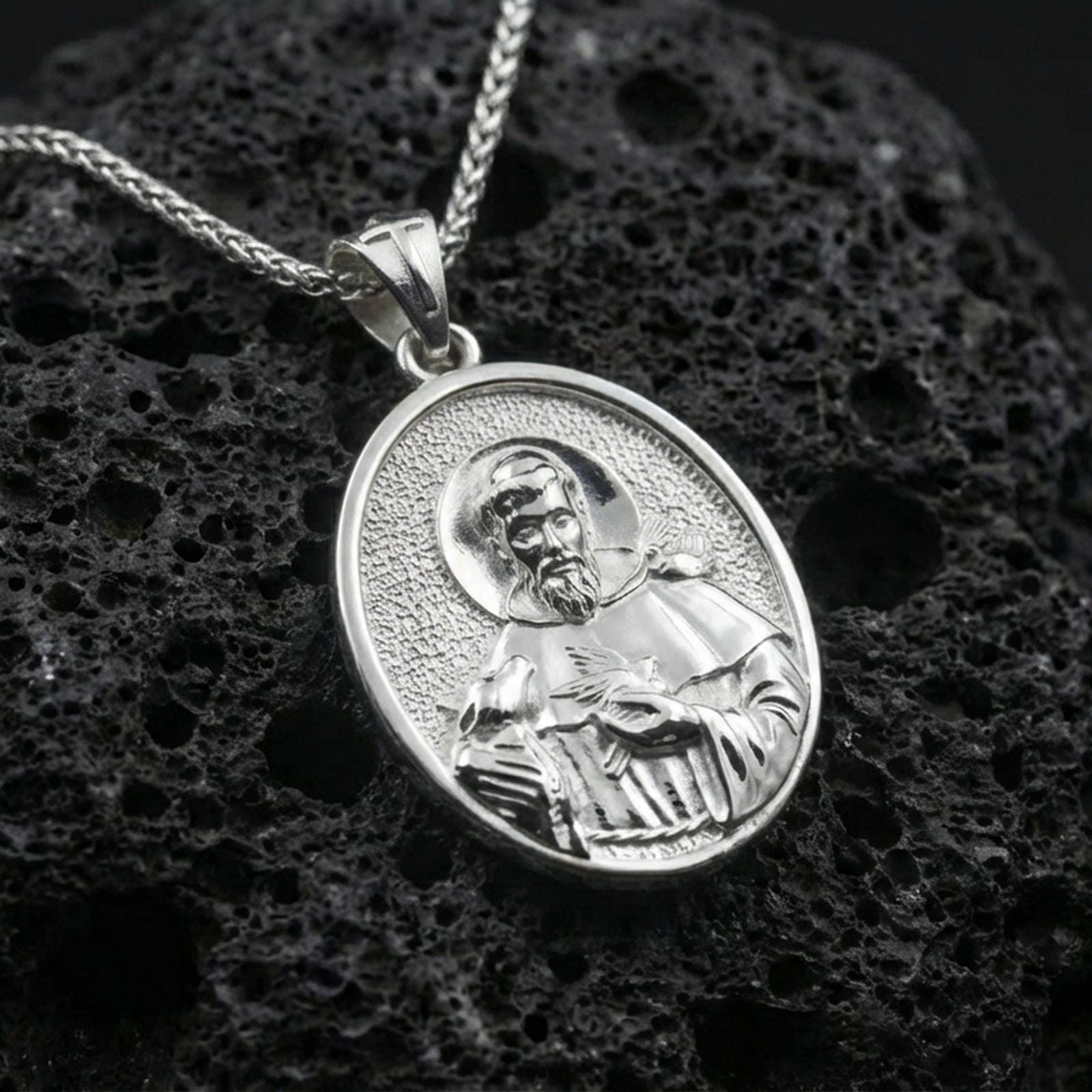 Polished silver St Francis oval pendant necklace on dark volcanic stone, crisp saint portrait engraving and smooth bezel edge, premium close-up
