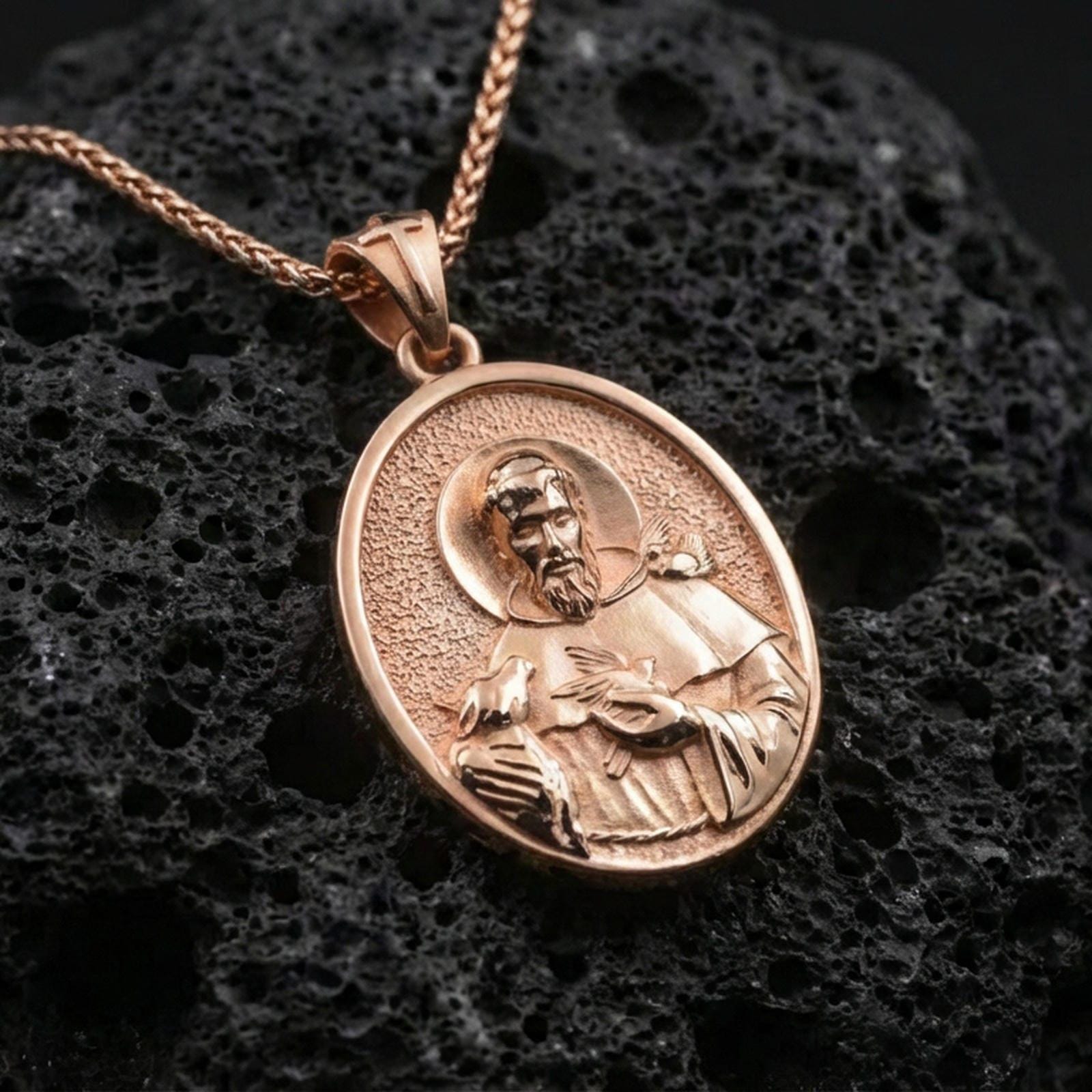 Rose gold plated St Francis oval pendant on matte volcanic stone, intricate saint engraving with bird and halo, sharp macro focus