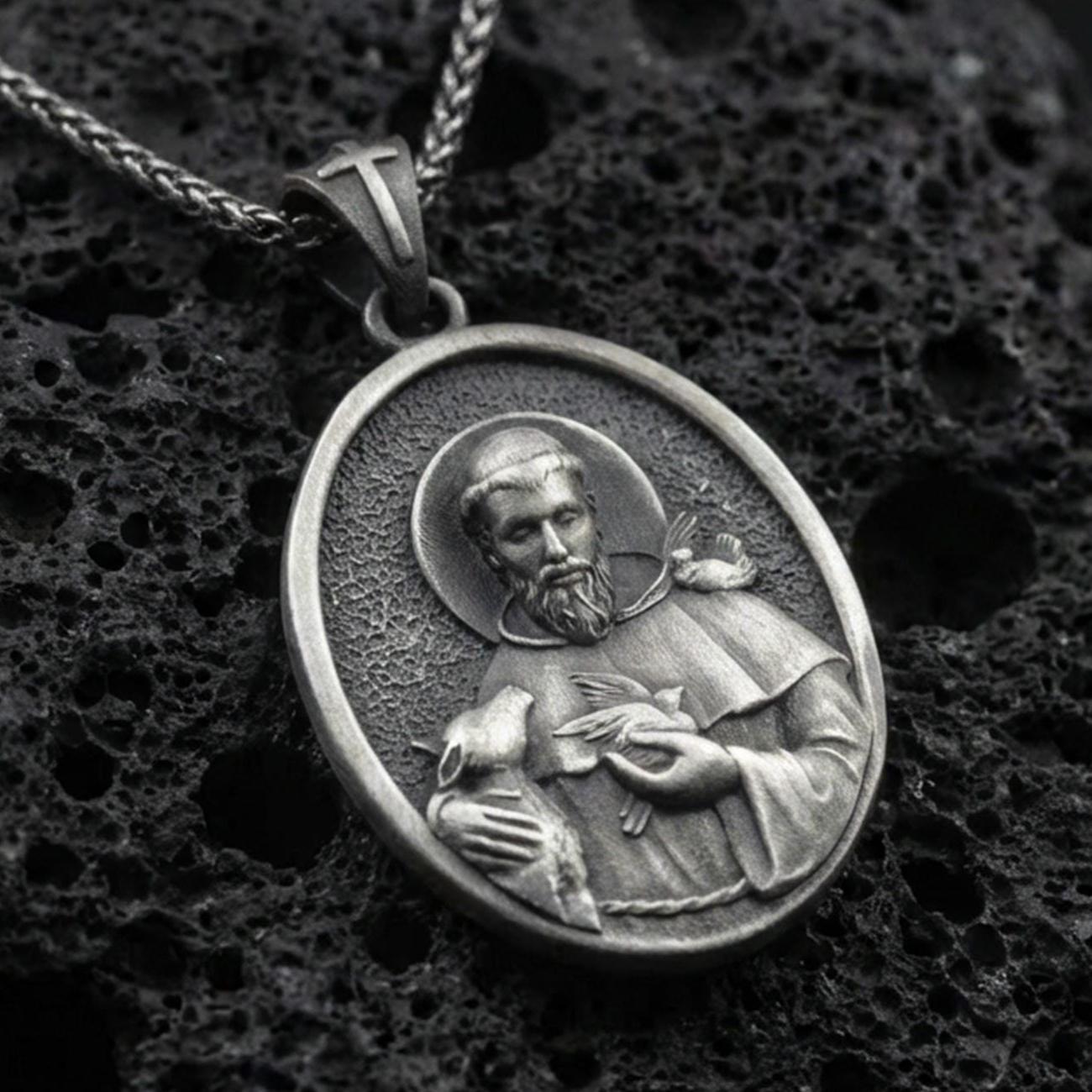 Oxidized sterling silver Saint Francis pendant on textured volcanic rock, oval medallion with halo relief and bird detail, macro studio shot