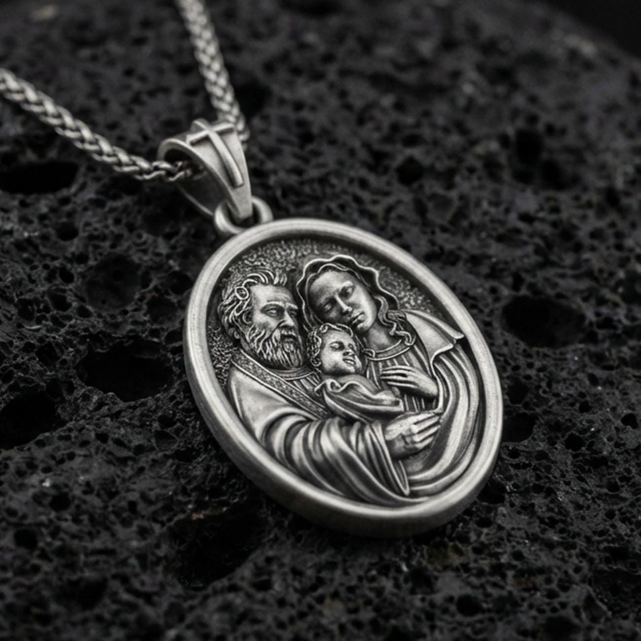 Oxidized sterling silver Holy Family oval medal on textured volcanic rock, detailed Mary Joseph and Child relief, macro studio shot