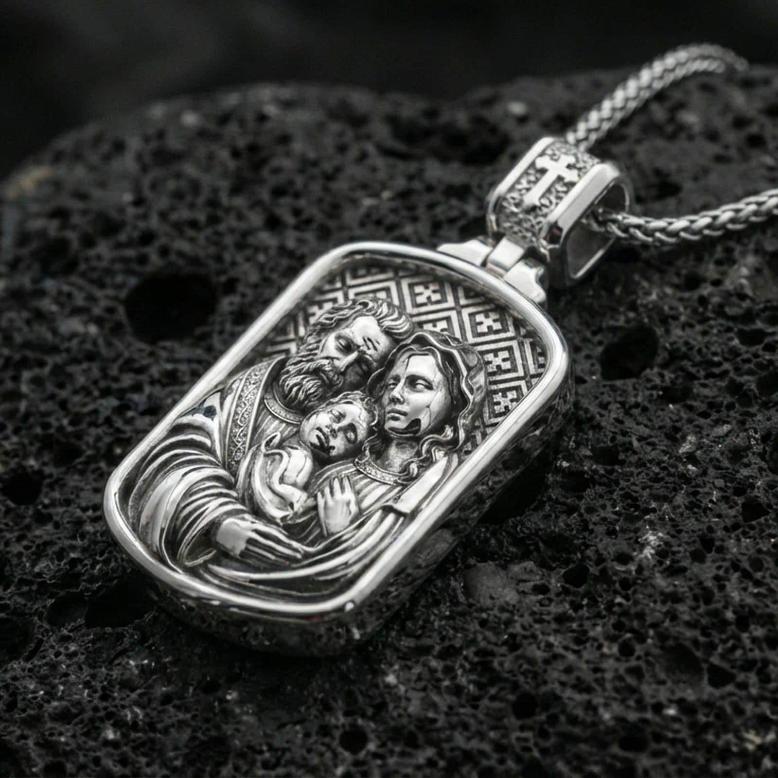 Polished silver Holy Family necklace pendant on dark volcanic stone, crisp engraved faces and geometric patterned background, premium close-up