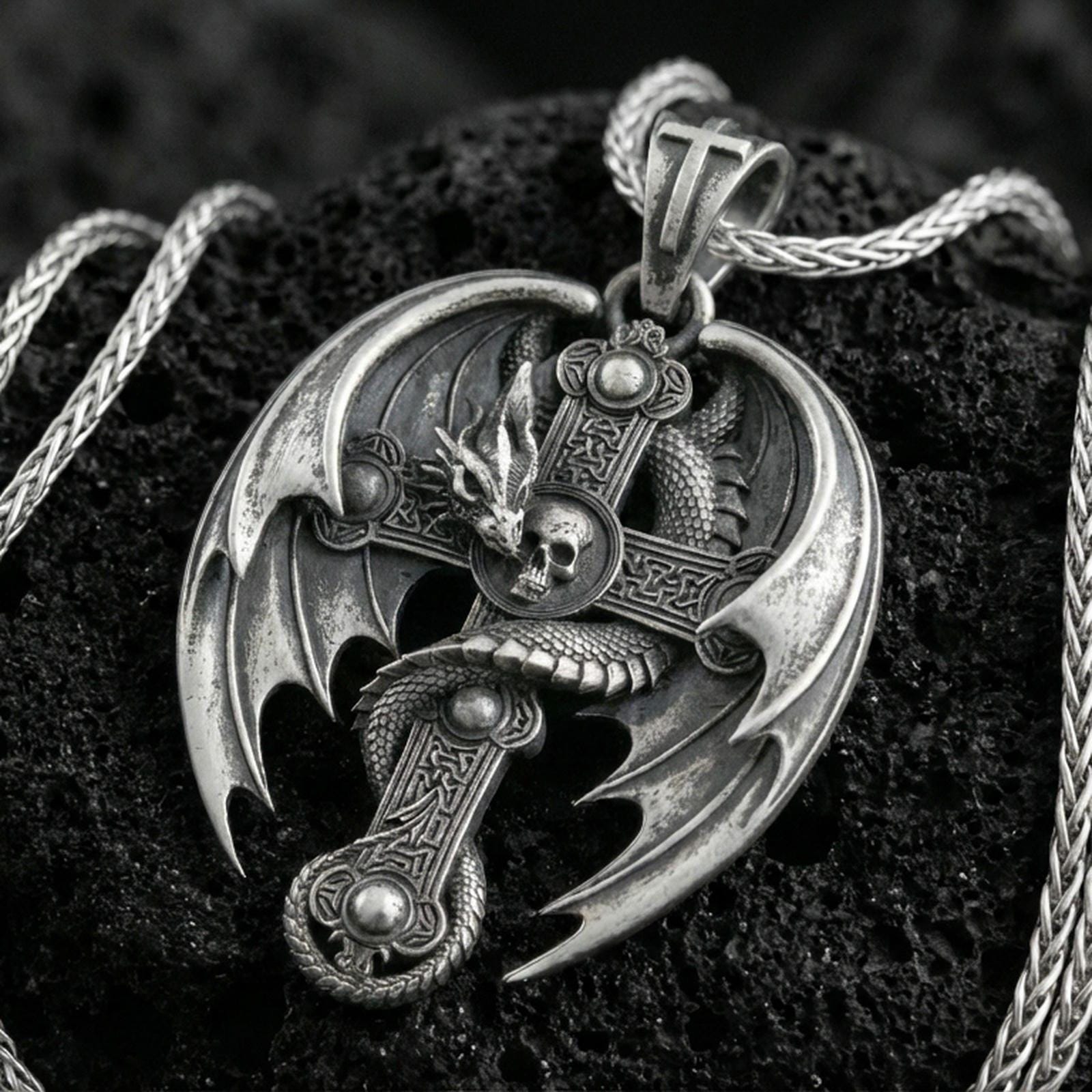 Oxidized sterling silver dragon cross pendant on textured volcanic rock, gothic winged dragon wrapped around medieval cross with skull detail, macro studio shot