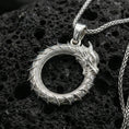Load image into Gallery viewer, Polished silver ouroboros dragon circle pendant on porous volcanic stone, detailed scale texture and horned head, product photo

