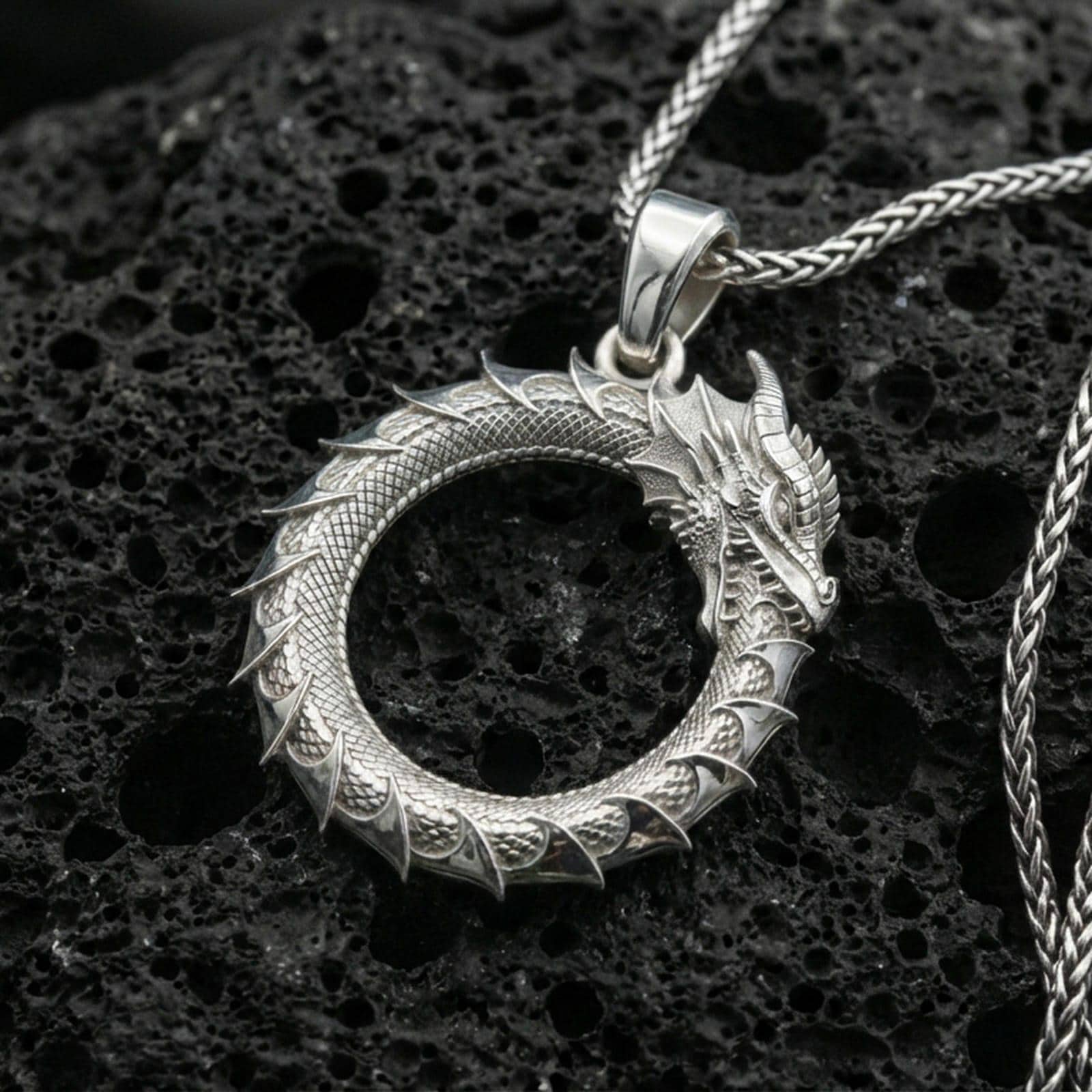 Polished silver ouroboros dragon circle pendant on porous volcanic stone, detailed scale texture and horned head, product photo