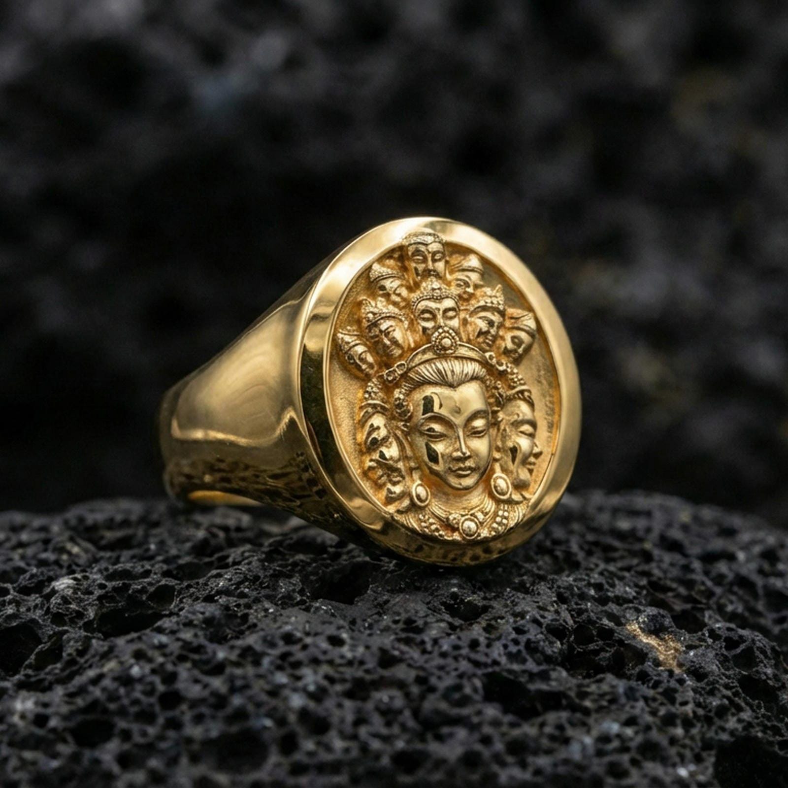 Gold plated multi-faced deity signet ring on dark volcanic rock, raised spiritual relief carving with ornate facial details