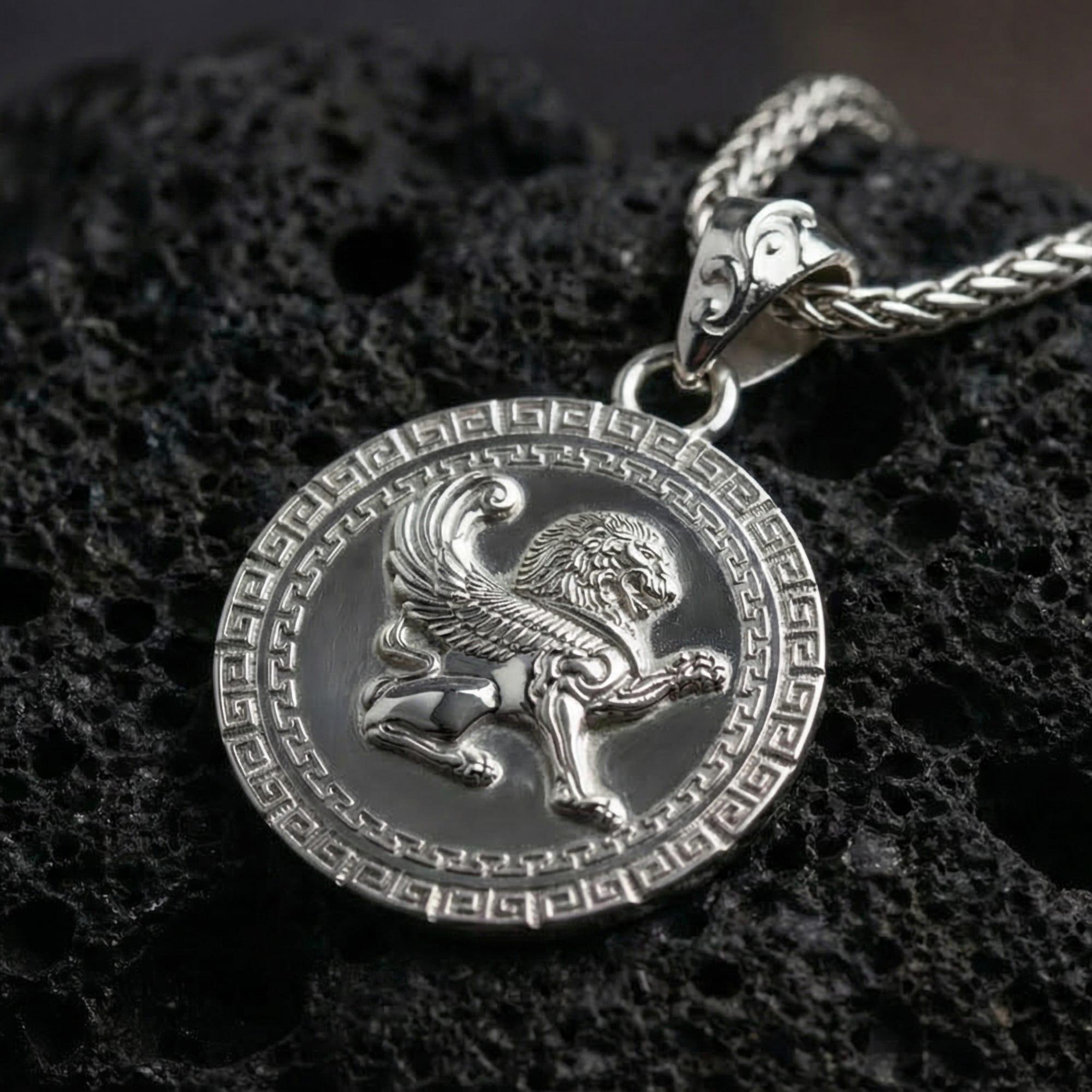 Polished silver winged lion pendant necklace photographed on volcanic rock with detailed mythical griffin style relief and Greek key border