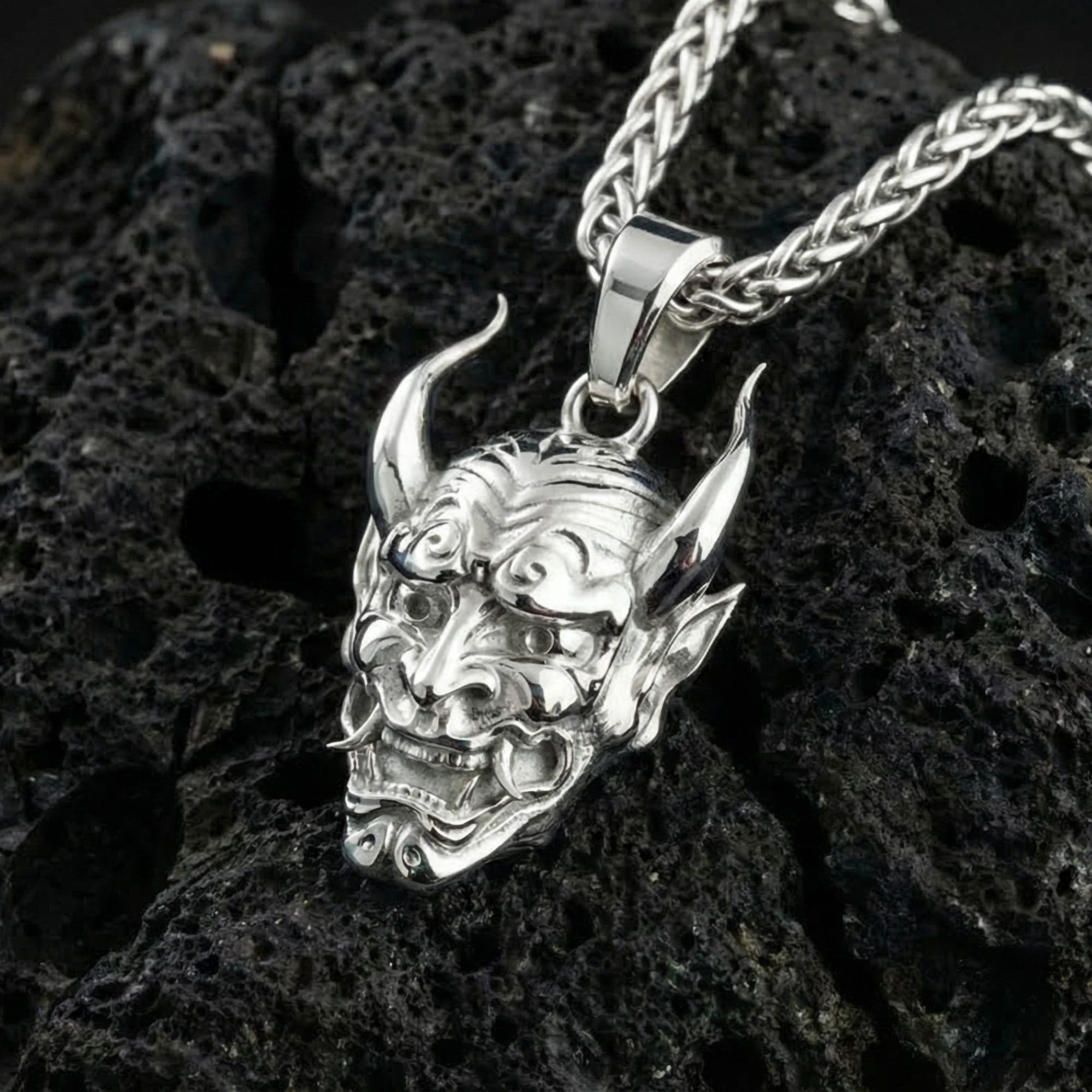 Polished silver Japanese oni mask pendant displayed on volcanic stone background with carved samurai spirit features