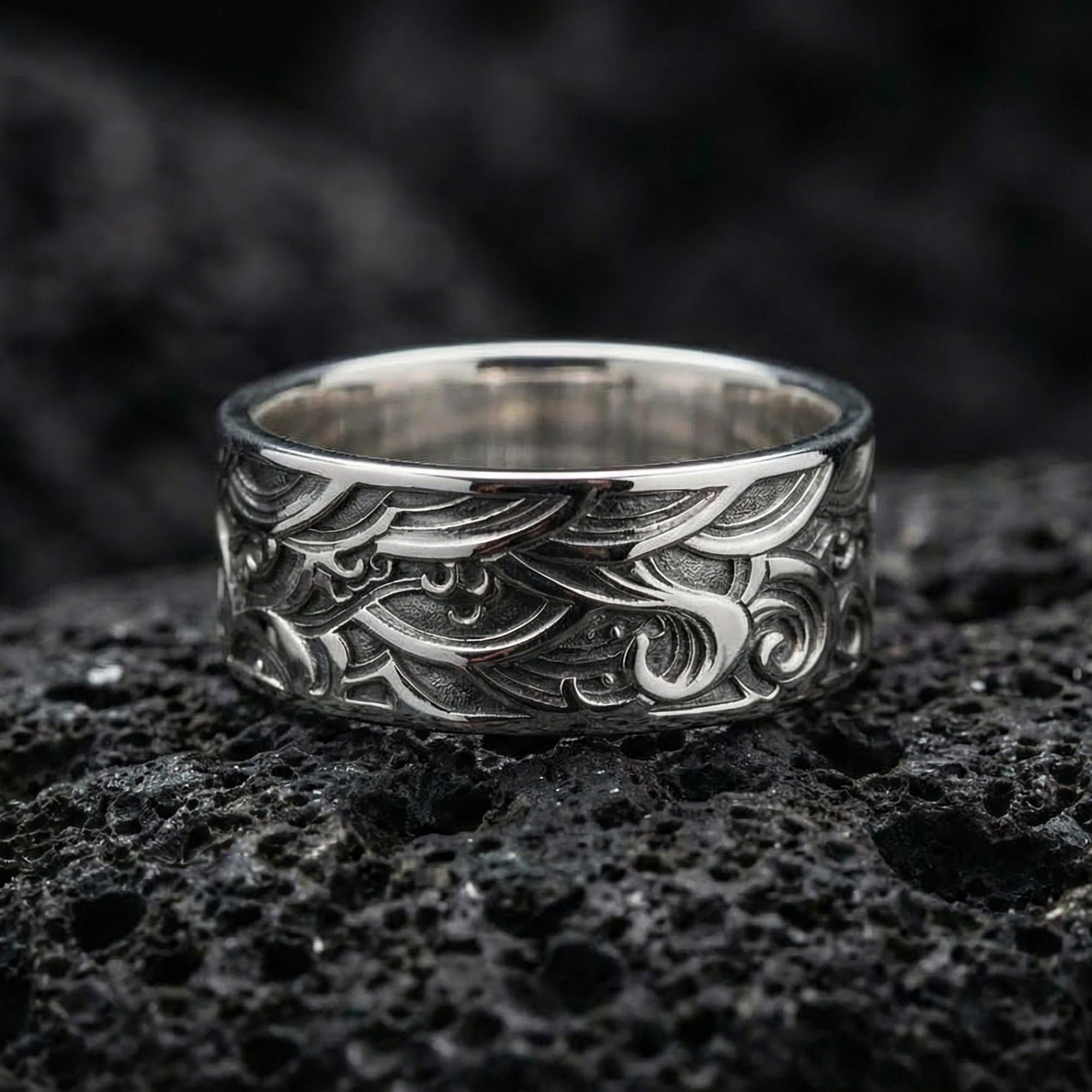 Polished silver wave engraved wedding band displayed on volcanic stone background, highlighting crisp scrollwork lines and clean reflective edges