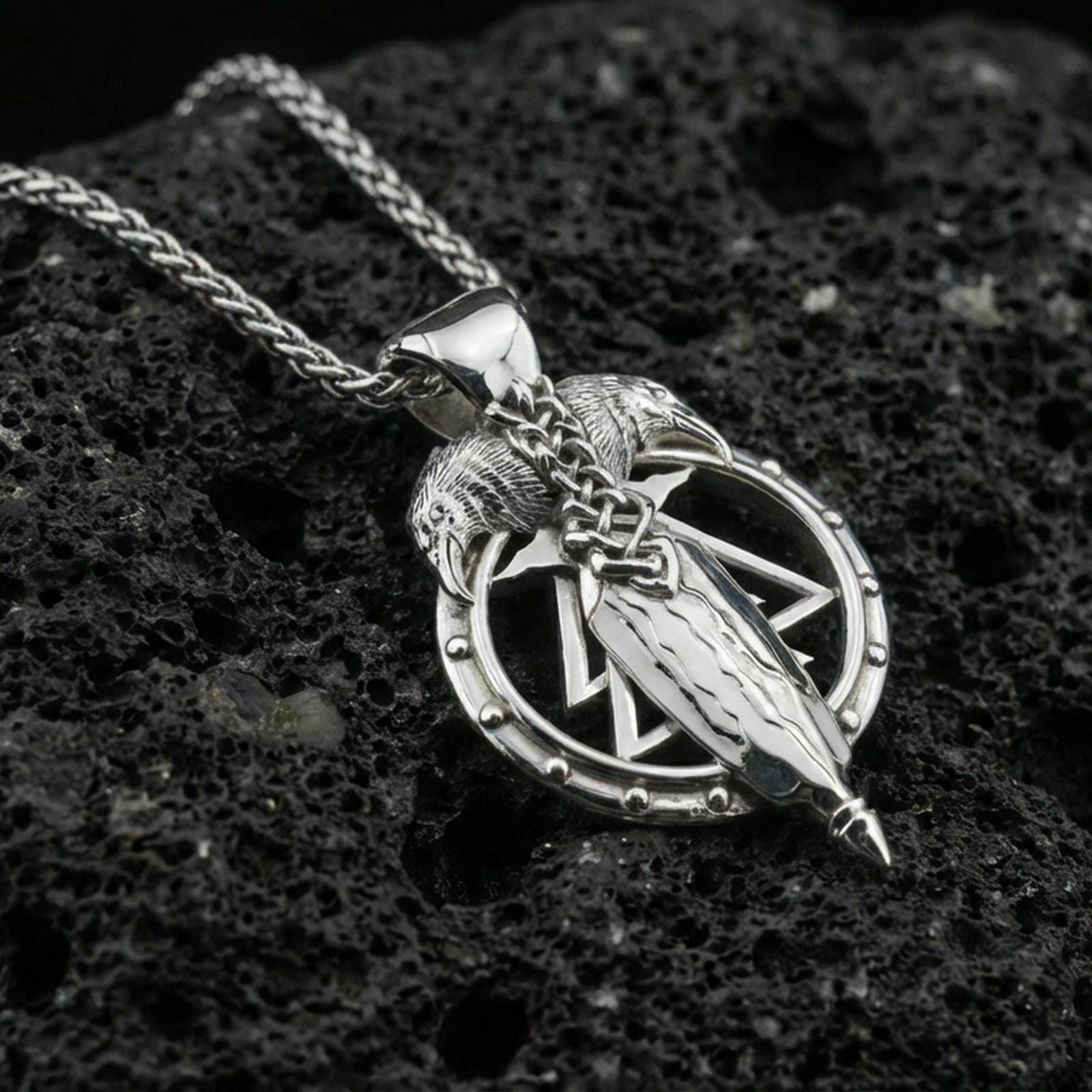 Polished silver raven and spear pendant on volcanic rock, sharp geometric rune-like frame detail, close-up studio lighting