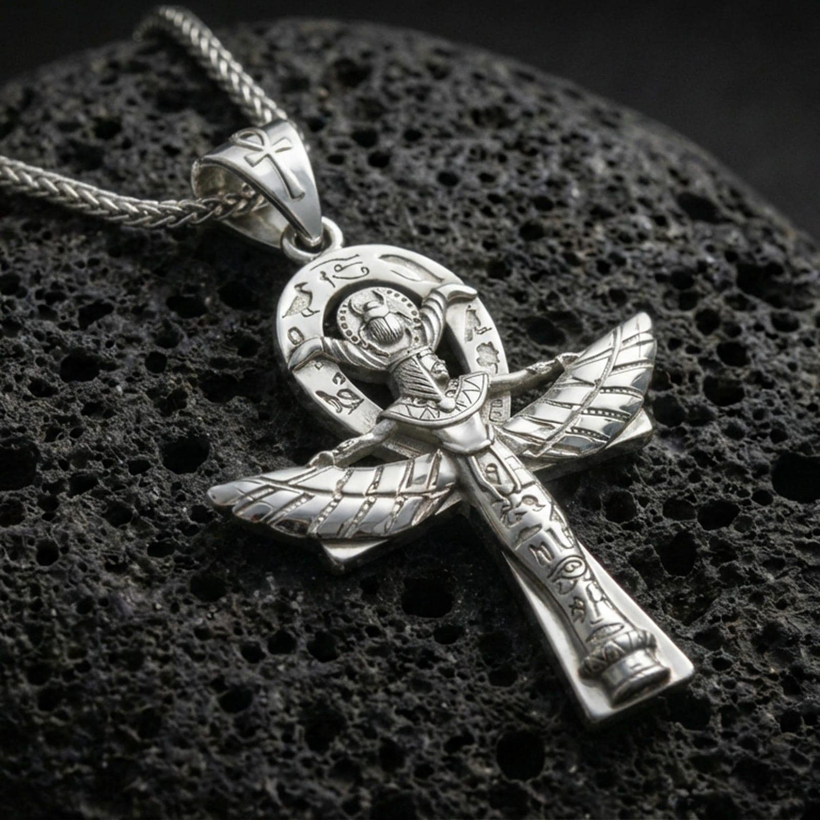Polished silver winged ankh cross necklace on volcanic rock, crisp etching and raised relief wings, close-up lighting