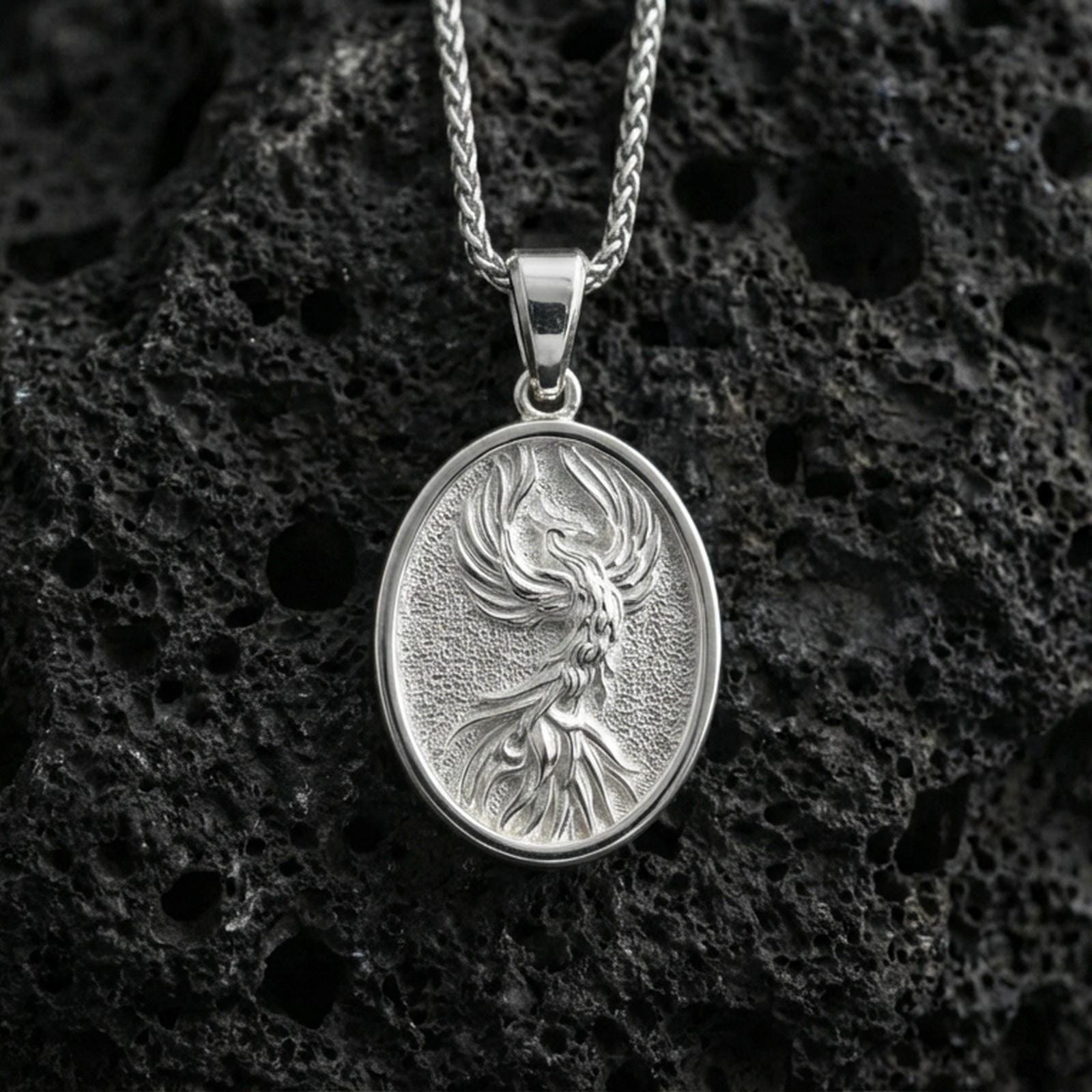 Polished silver phoenix medallion necklace on volcanic rock, crisp raised flame-feather relief and smooth oval frame, close-up