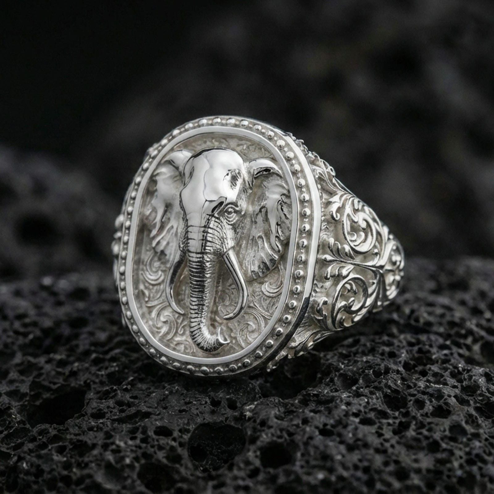 Polished silver elephant signet ring on volcanic rock, raised elephant head relief with beaded frame and scrollwork details, close-up