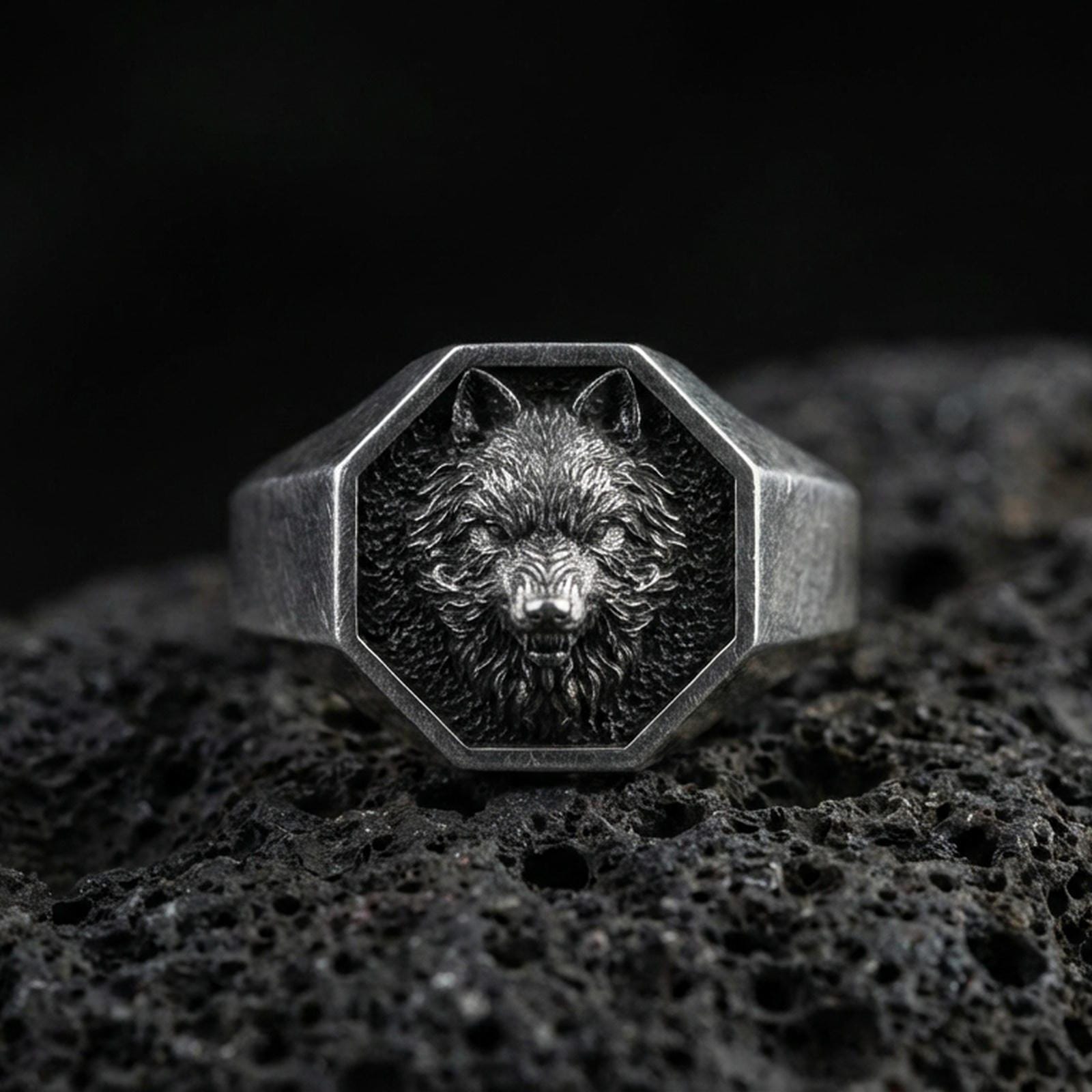 Oxidized sterling silver wolf signet ring on volcanic rock, octagonal face with raised wolf head relief and dark textured background, macro product photo