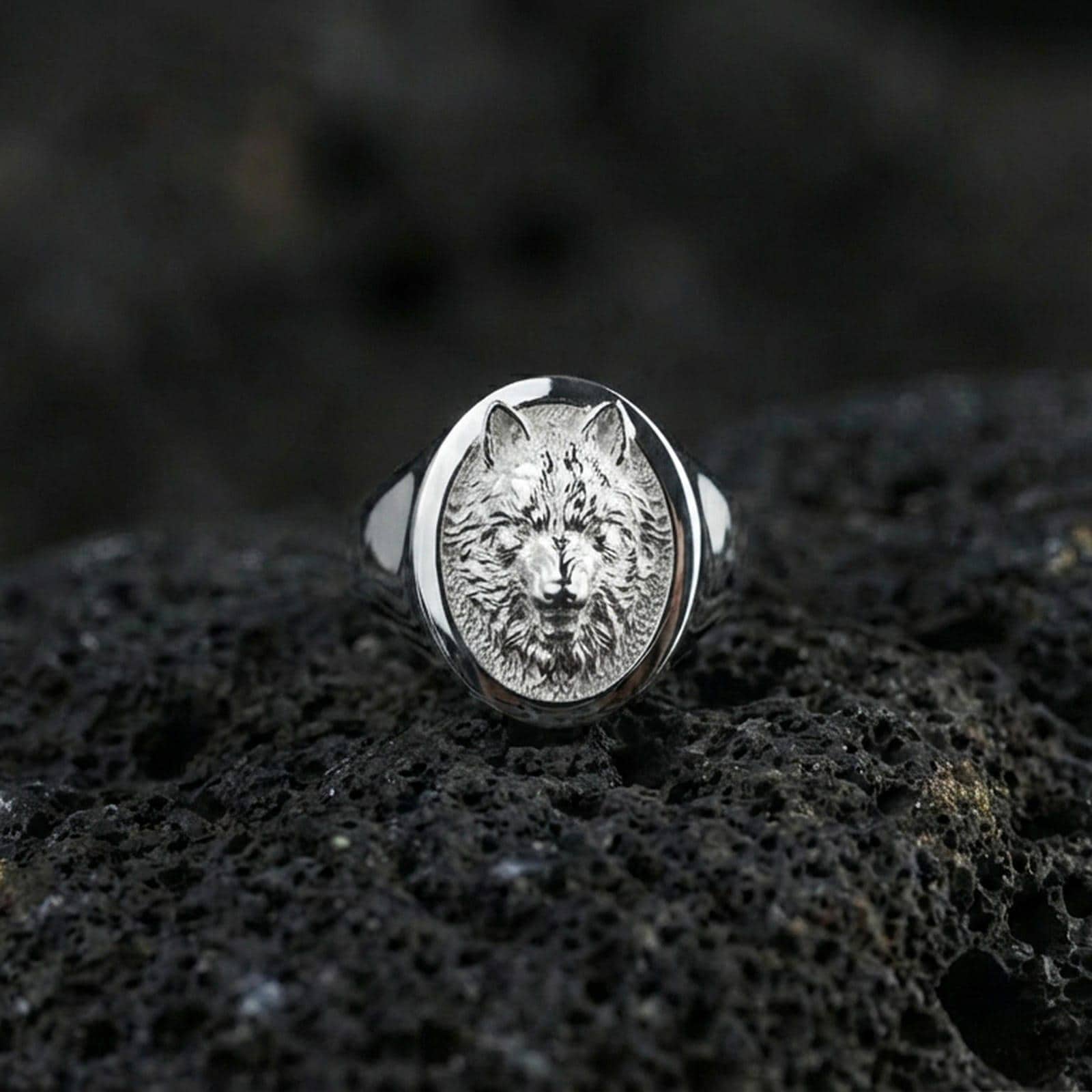 Polished silver wolf signet ring on volcanic rock, oval bezel framing a raised wolf portrait with sharp edge rim, premium close-up shot