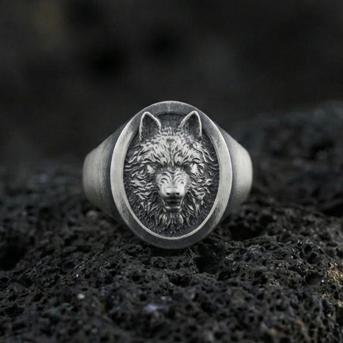Oxidized sterling silver wolf signet ring on volcanic rock, oval face with detailed wolf head relief and dark recessed background, macro jewelry photo