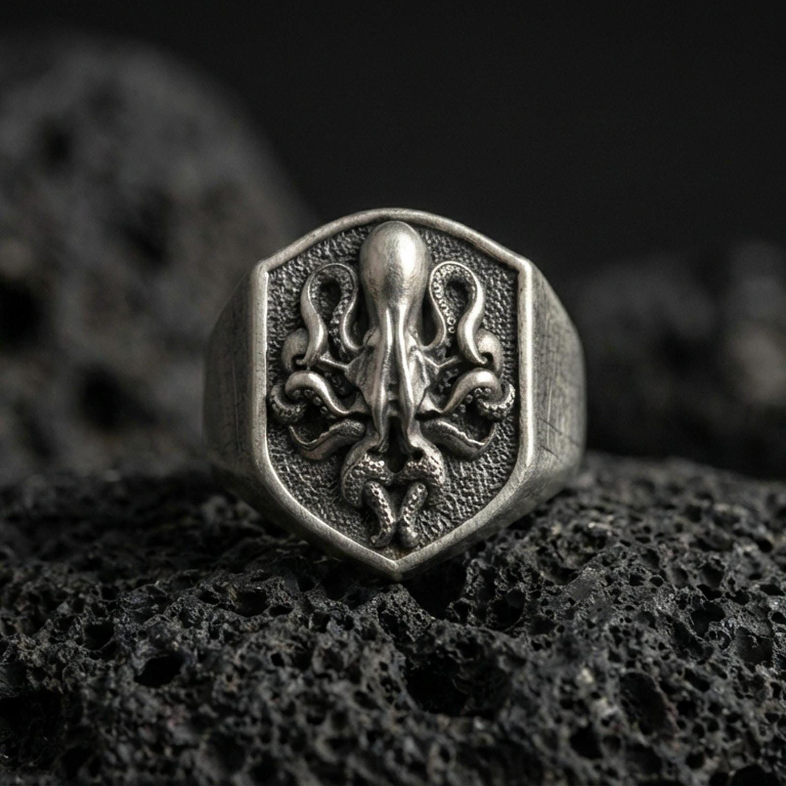 Oxidized sterling silver octopus signet ring on volcanic rock, shield-shaped face with raised octopus emblem and dark textured background, macro jewelry photo