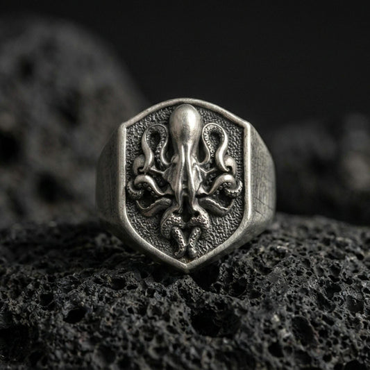 Oxidized sterling silver octopus signet ring on volcanic rock, shield-shaped face with raised octopus emblem and dark textured background, macro jewelry photo