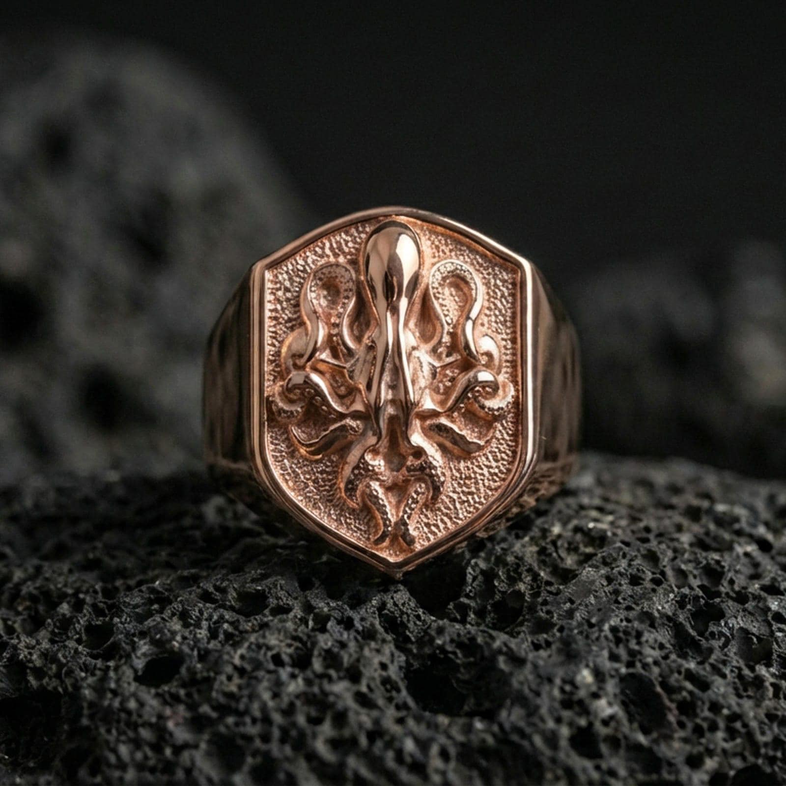 Rose gold plated octopus signet ring on volcanic rock, shield octopus crest signet with sculpted tentacles and hammered-look recessed surface, luxury product photo