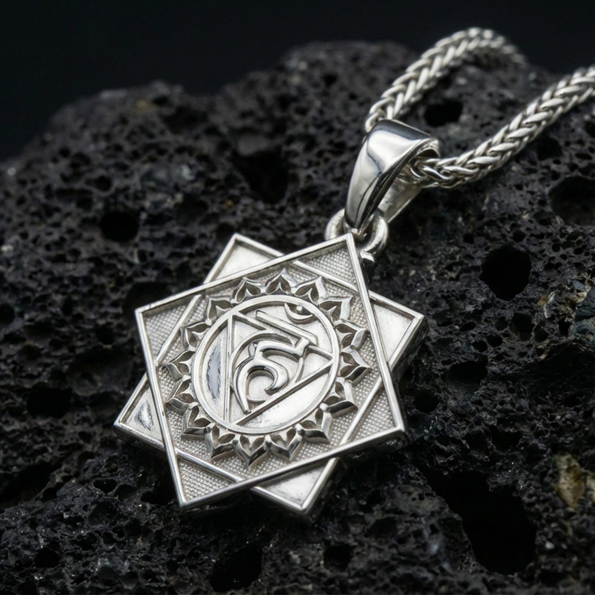 Polished silver Vishuddha chakra necklace pendant on black volcanic stone background, crisp engraved lines, high-end product shot