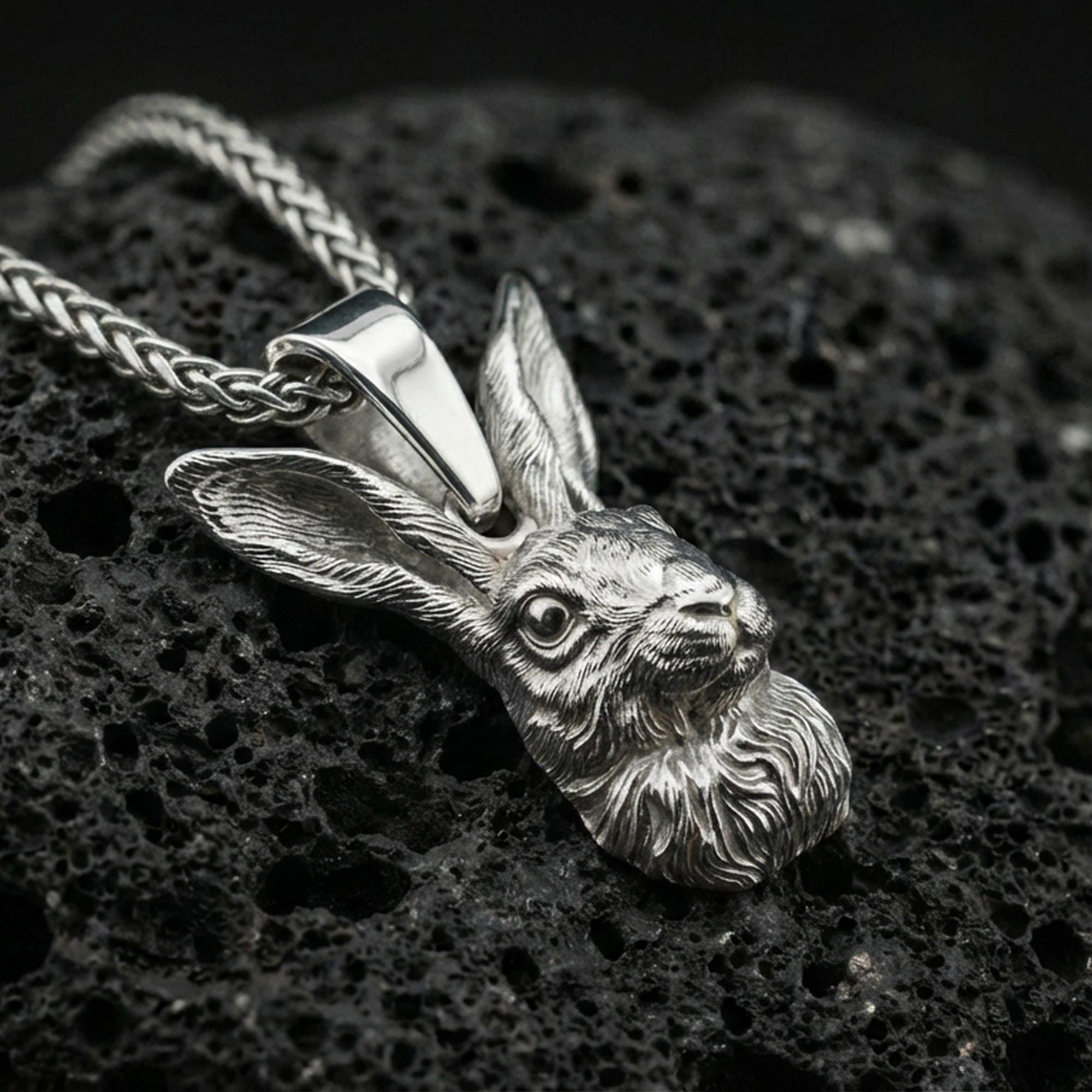 Polished silver rabbit necklace pendant on black volcanic stone background, crisp sculpted hare face detail, high-end product shot