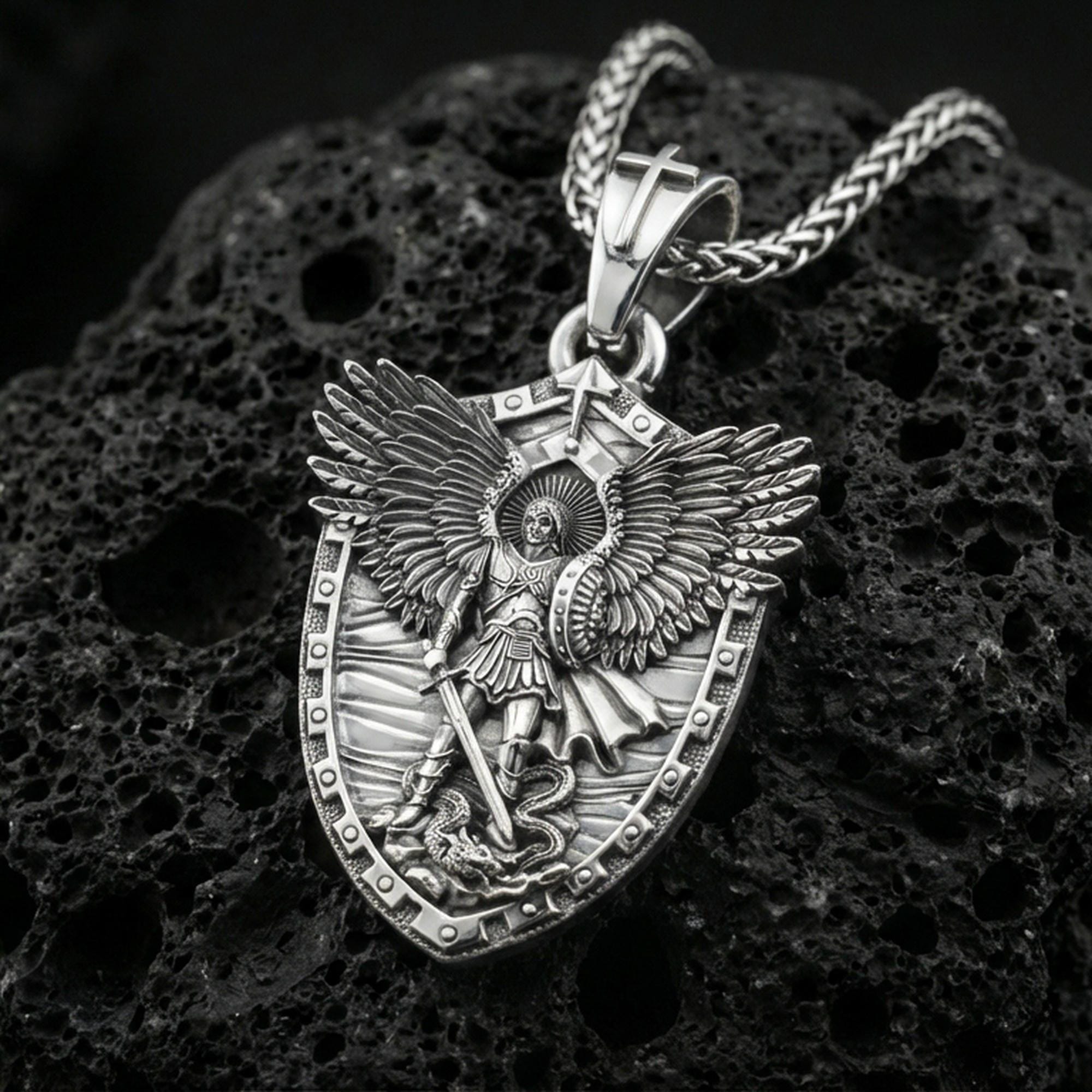 Polished silver Archangel Michael necklace pendant on black volcanic stone background, crisp shield border and angel figure, high-end product shot