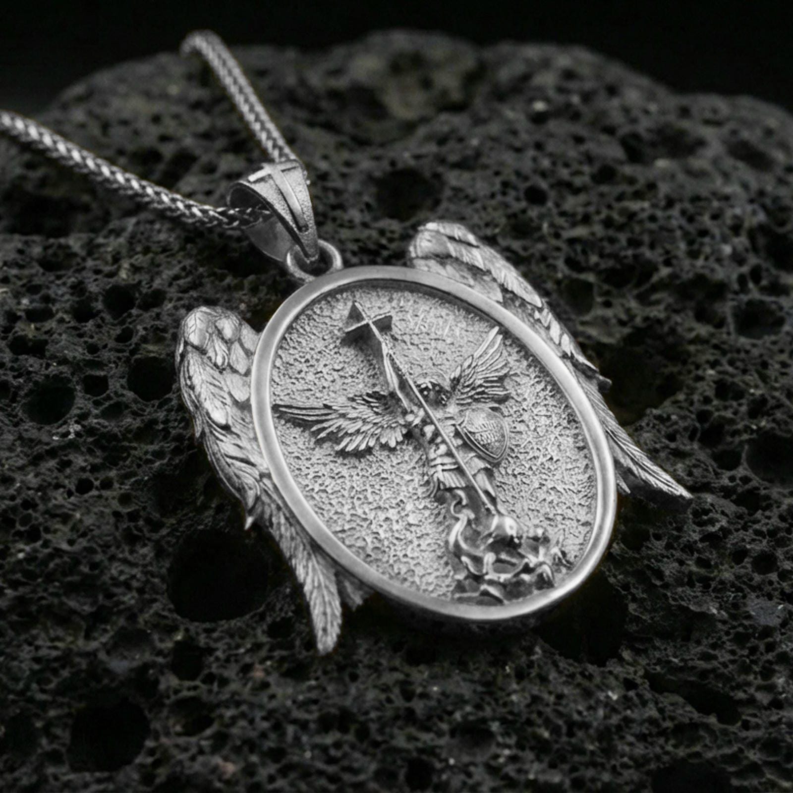 Polished silver Saint Michael necklace pendant on black volcanic stone background, crisp archangel relief with spear and shield, high-end product shot