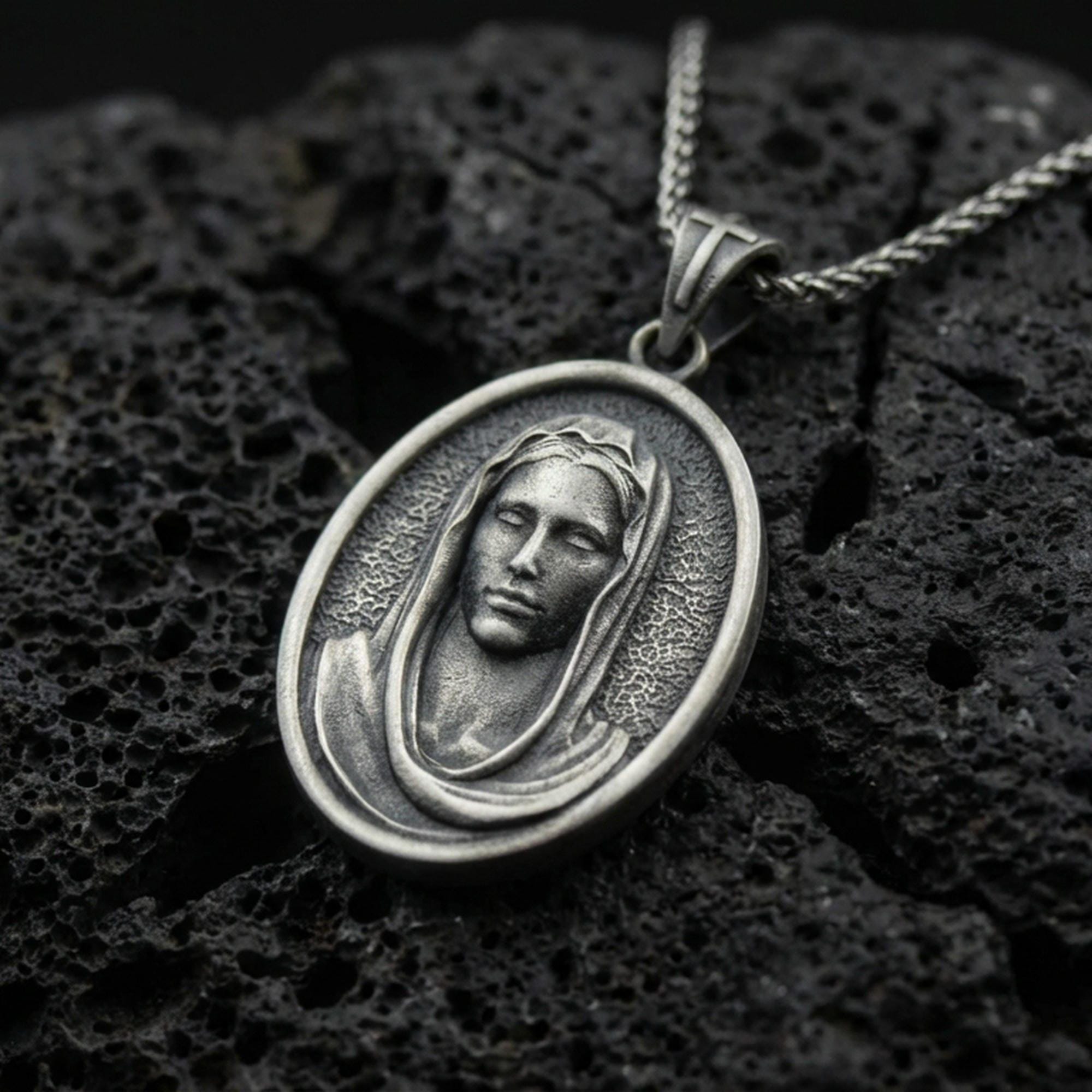 Oxidized Veiled Madonna pendant on textured volcanic rock, macro close-up, serene Virgin Mary relief detail, premium studio lighting