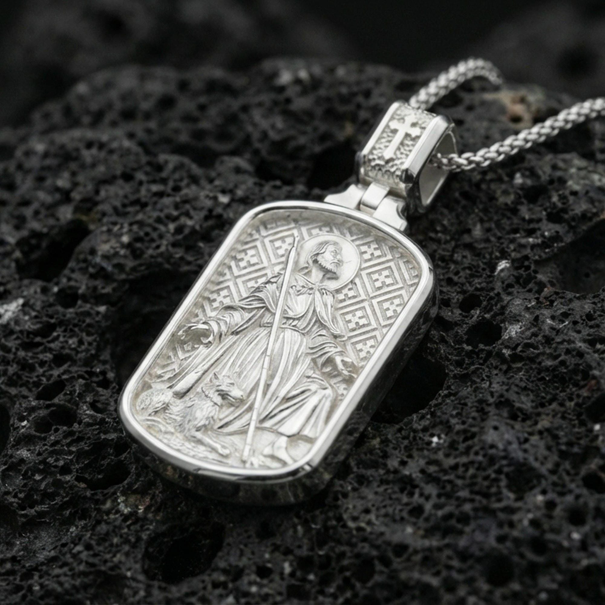 Polished silver Saint Roch dogtag necklace pendant on black volcanic stone background, crisp rectangular frame and patterned backdrop, high-end product shot