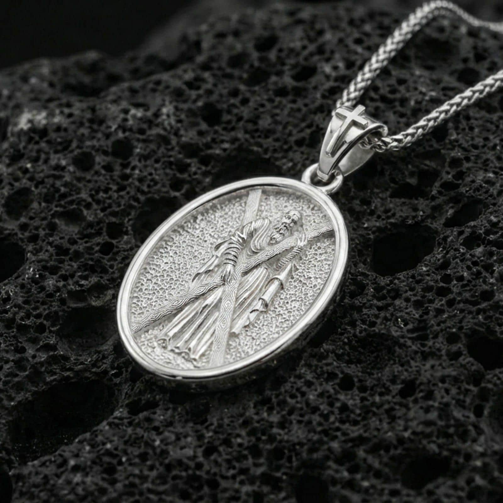 Polished silver Saint Andrew oval medallion pendant on black volcanic stone, crisp edge frame and textured background, luxury product photo
