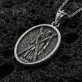Load image into Gallery viewer, Oxidized Saint Andrew oval pendant necklace on volcanic rock, apostle carrying cross relief, macro close-up, premium studio lighting
