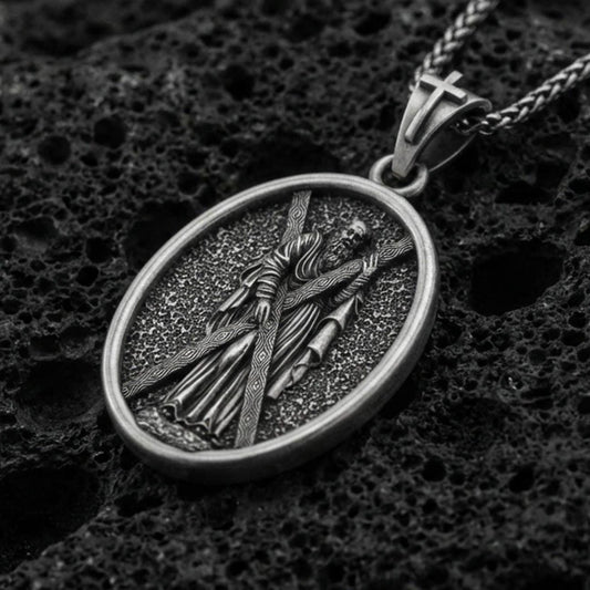 Oxidized Saint Andrew oval pendant necklace on volcanic rock, apostle carrying cross relief, macro close-up, premium studio lighting