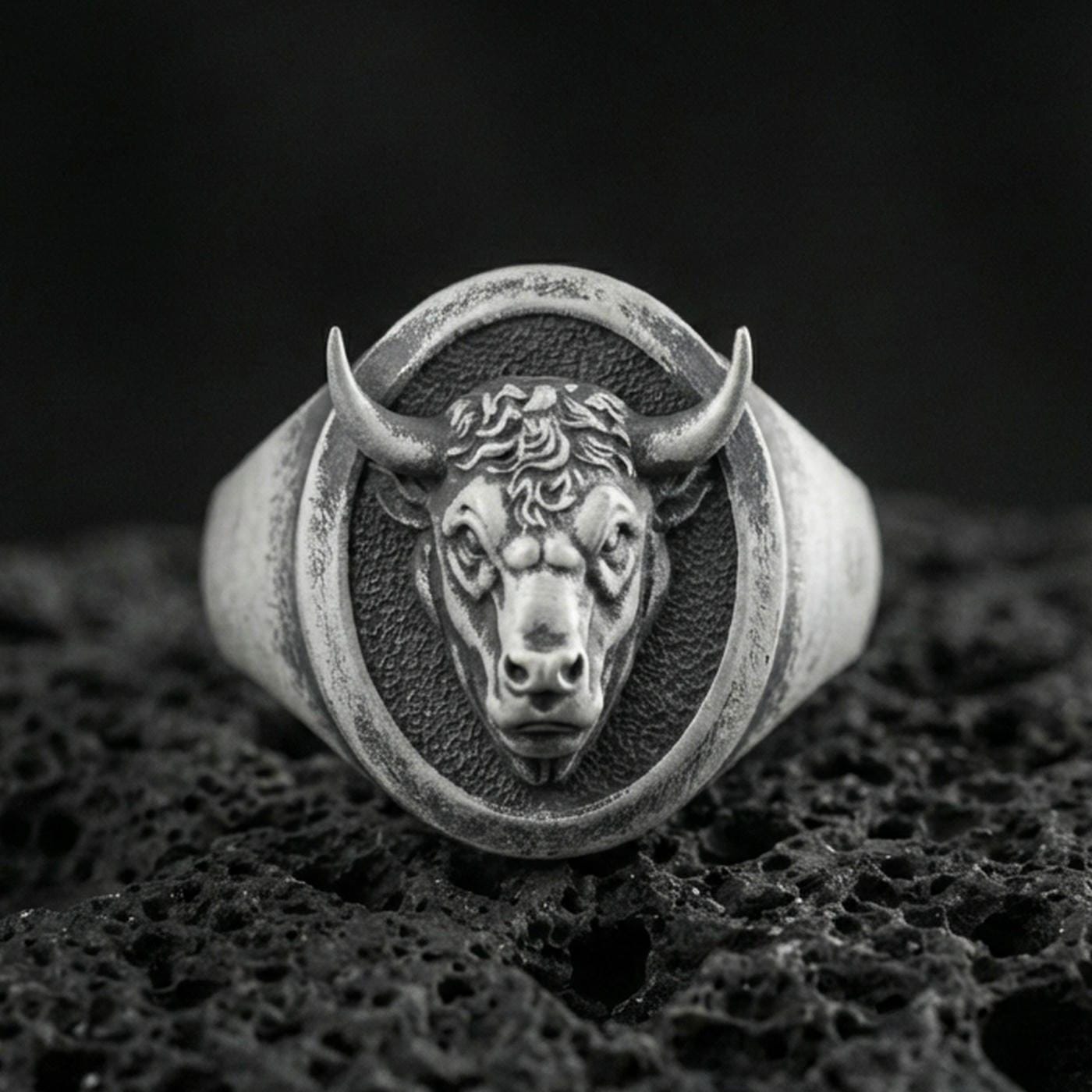 Oxidized sterling silver bull signet ring on textured volcanic rock, front-facing horned head relief with oval face in macro studio shot