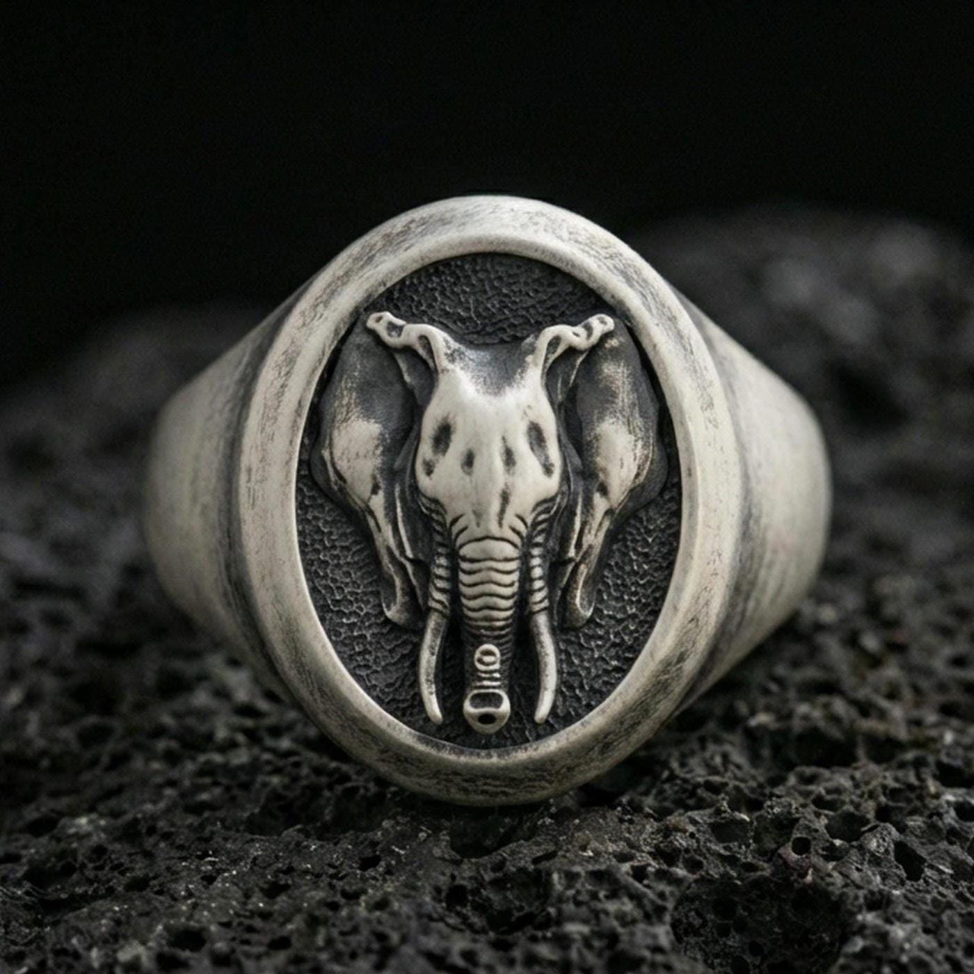 Oxidized sterling silver elephant signet ring on textured volcanic rock, macro close-up showing oval face with frontal elephant head, tusks, and trunk detail