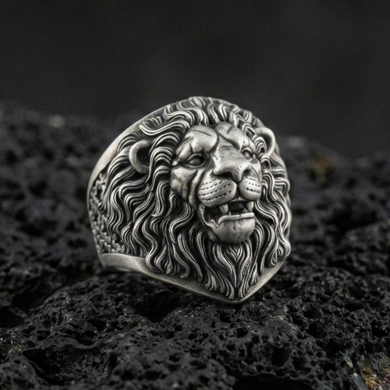 Oxidized sterling silver lion head ring on textured volcanic rock, macro close-up showing roaring expression, detailed mane curls, and sculpted fangs