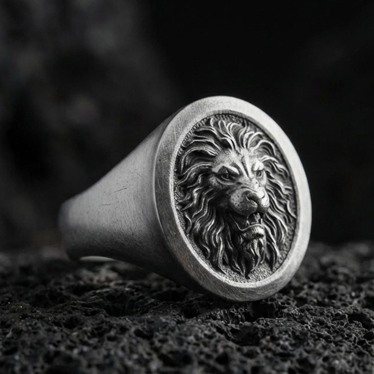 Oxidized sterling silver lion signet ring on textured volcanic rock, macro close-up highlighting the roaring lion head relief and dark recessed background