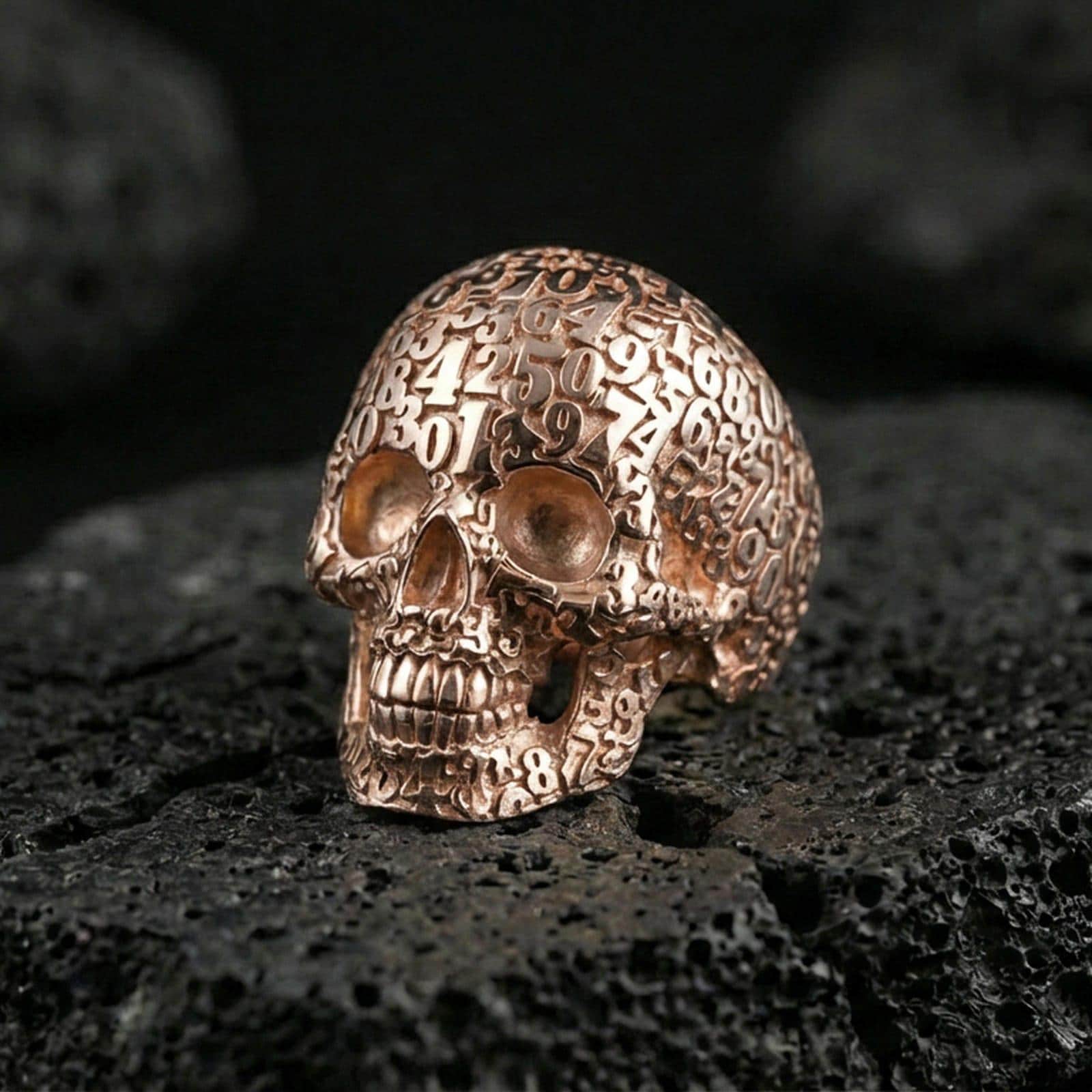 Cronus skull numerology ring in a rose gold plated finish on volcanic rock, ornate number-covered skull detail, luxury studio close-up