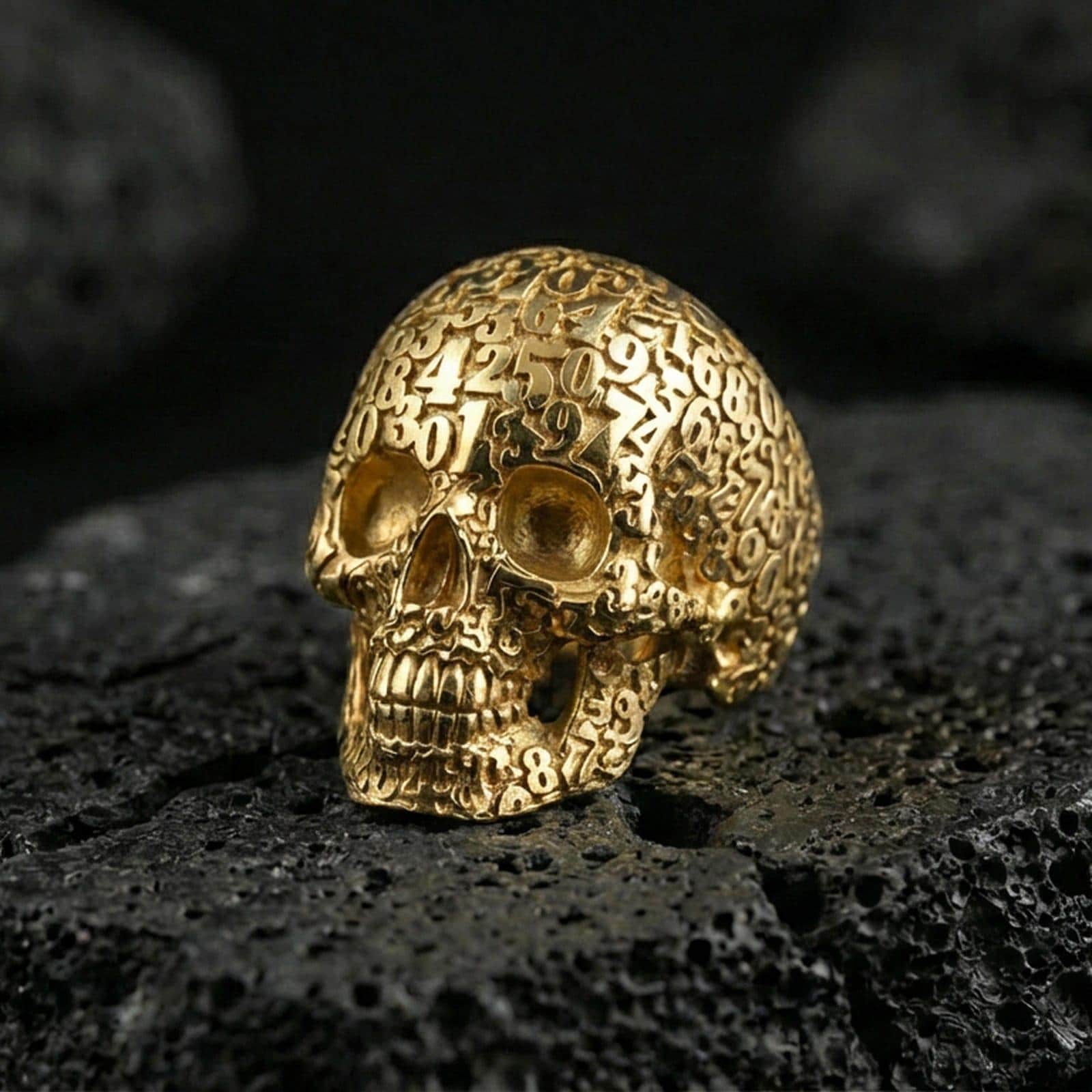 Numerology skull signet ring in a gold plated finish on volcanic rock, bold memento mori skull covered with engraved numbers, premium macro image