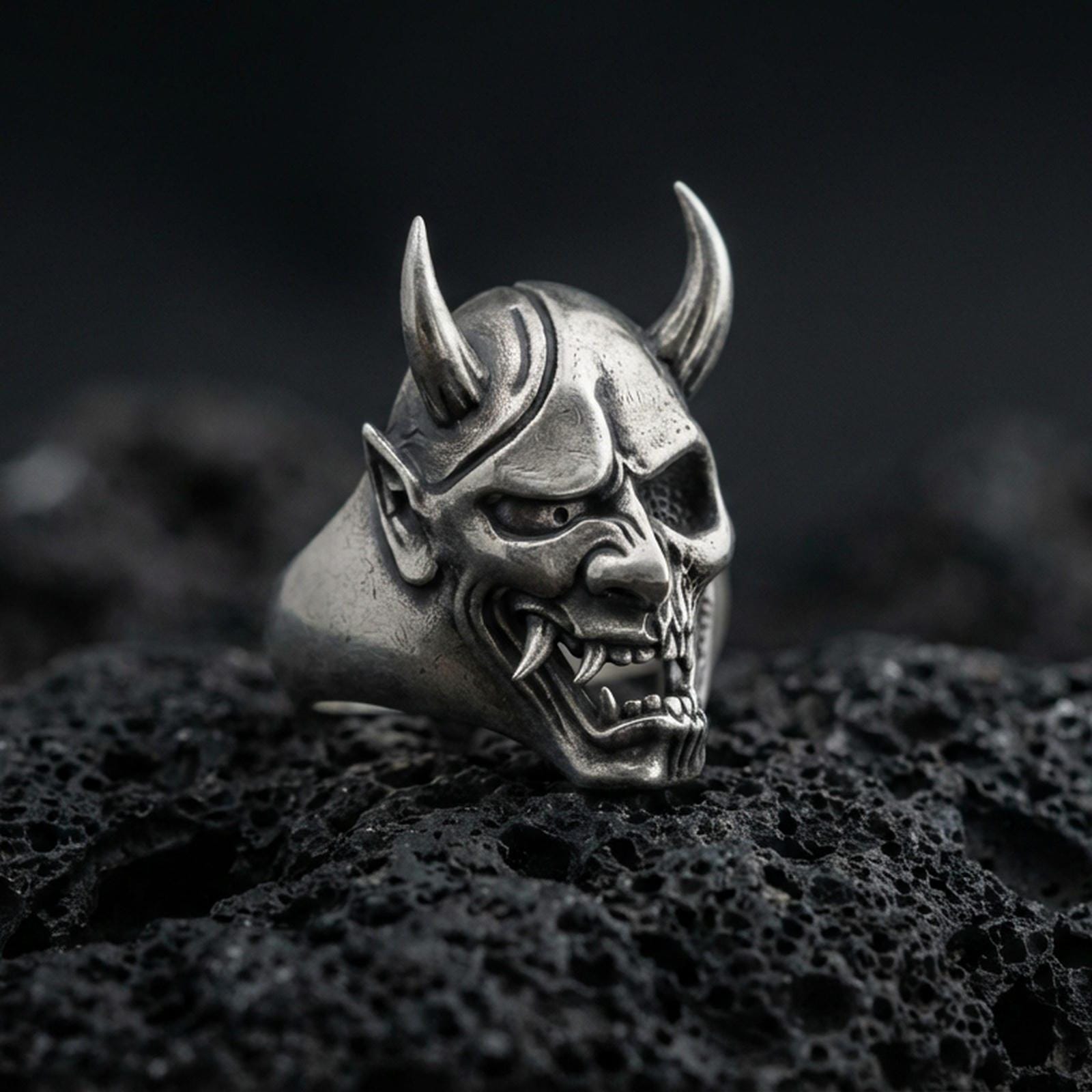 Sterling silver undead oni mask ring in an oxidized finish on volcanic rock, Japanese yokai demon face with horns, macro studio product shot
