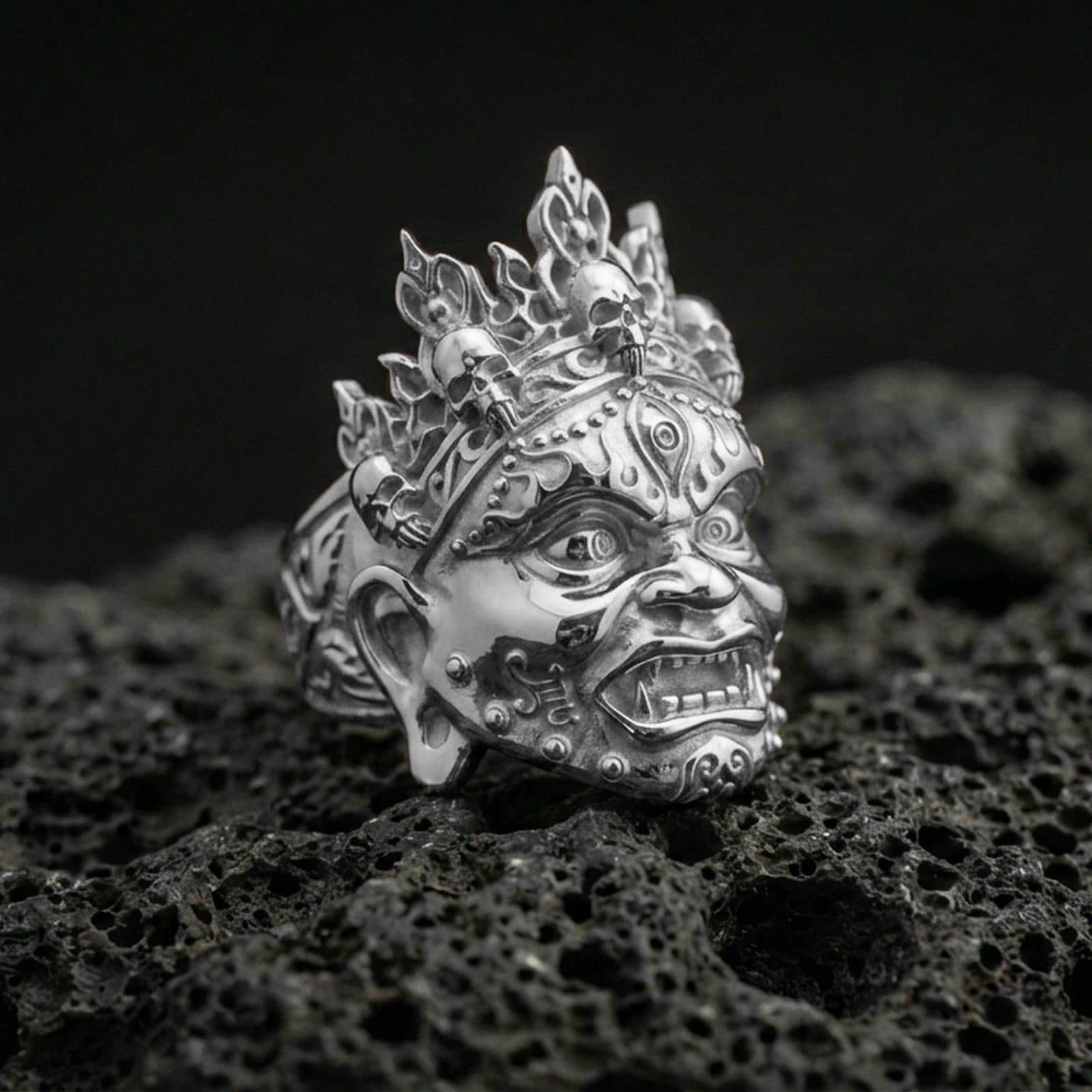 Yama god of death mask ring in polished silver finish on volcanic rock, sculpted fangs and ornate forehead engravings, premium studio close-up